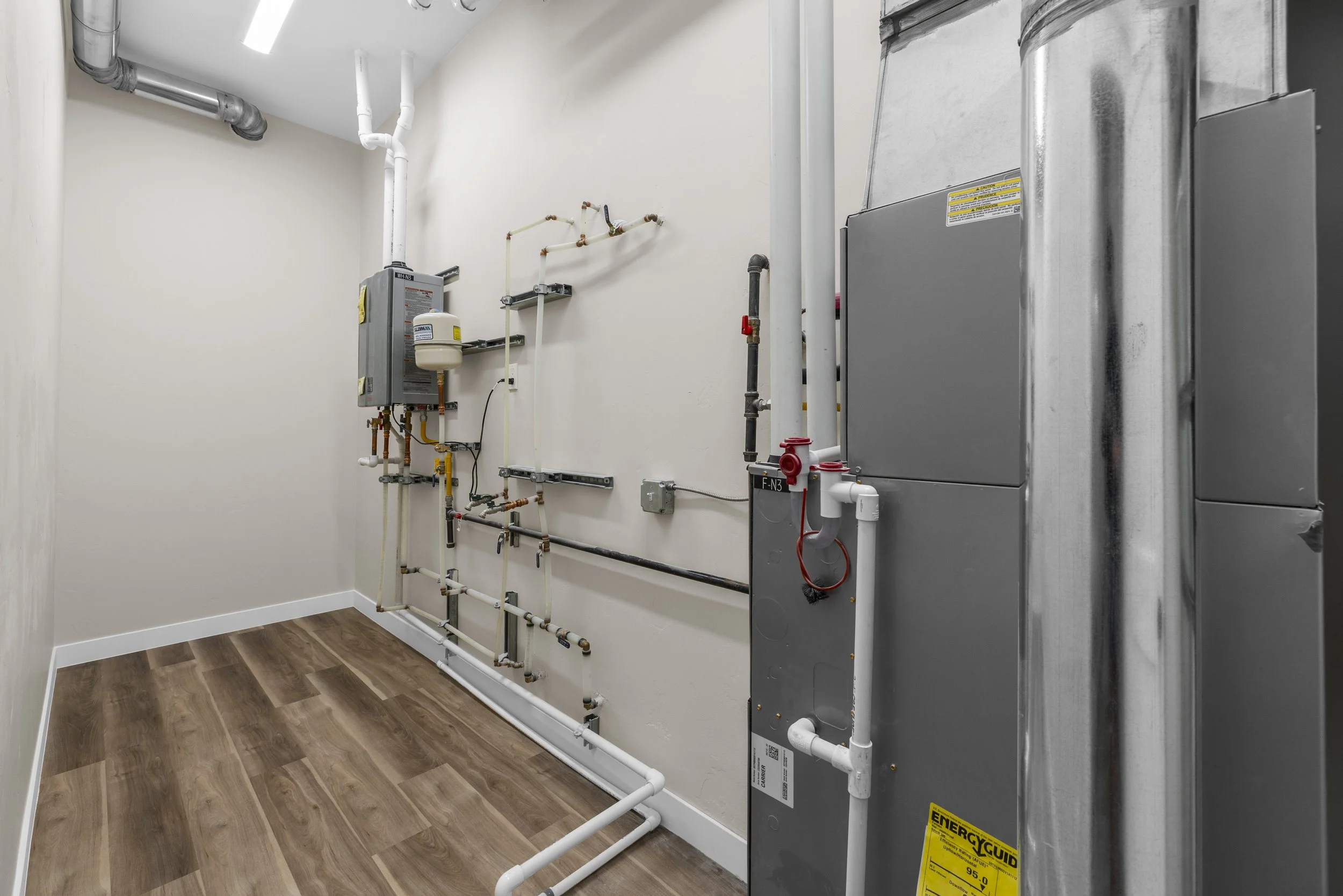 Empty utility room with HVAC and plumbing pipes, electrical components, and a heating system on a white wall, with wood flooring.