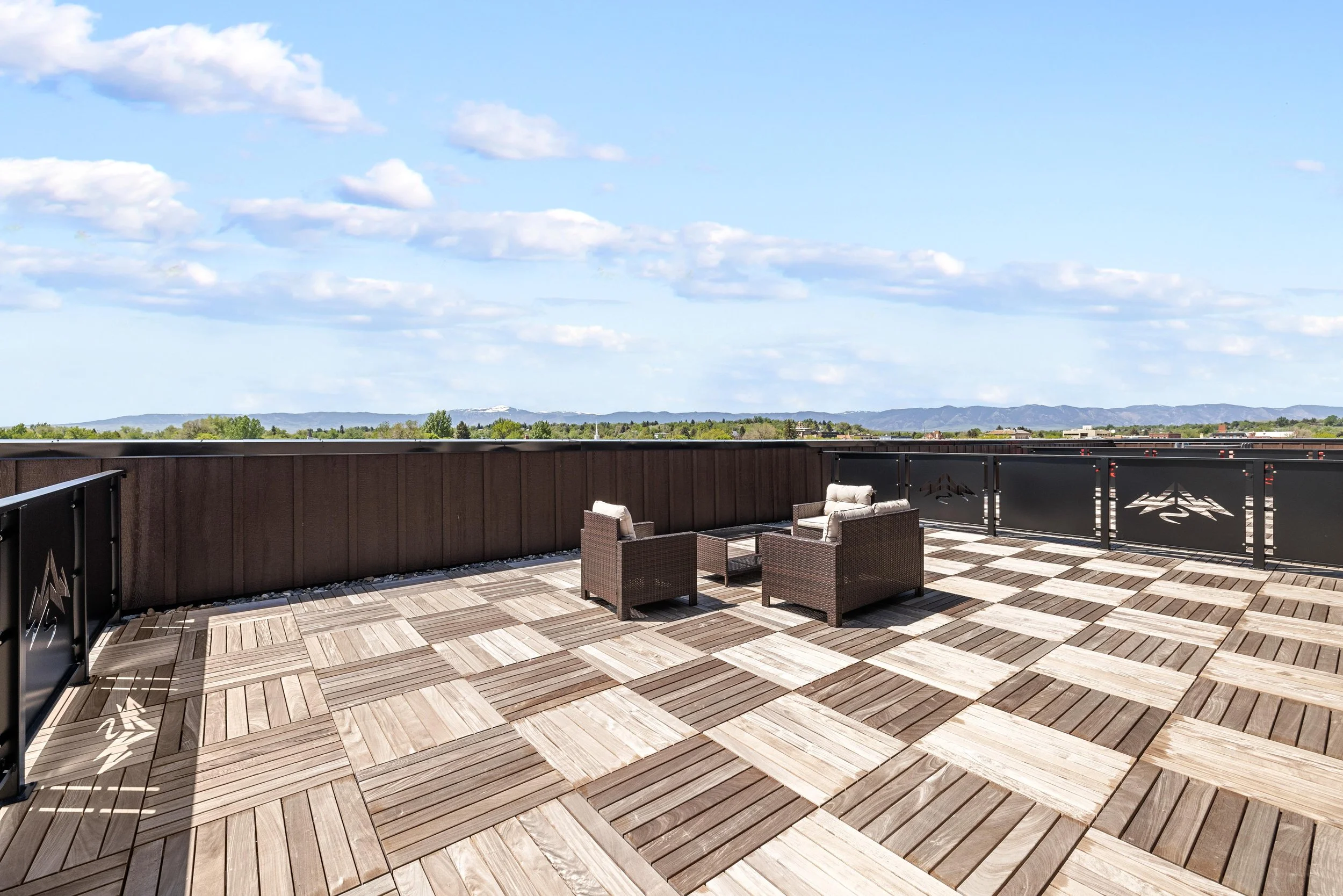 Rooftop balcony with outdoor seating, wooden tile flooring, brown railing, and distant mountain view under a partly cloudy sky.