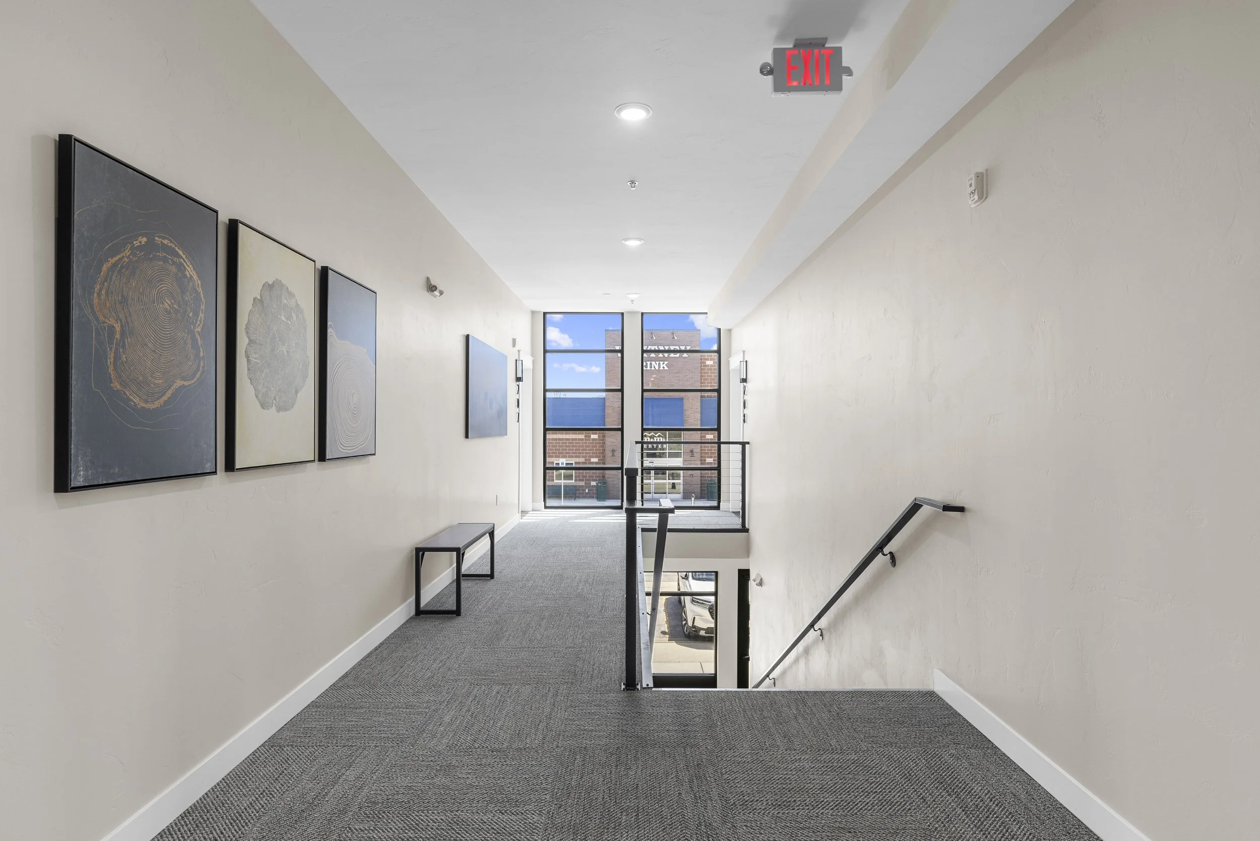 Hallway inside a modern building with artwork on the left wall, a bench, and a window at the end showing a parking lot and brick building outside.