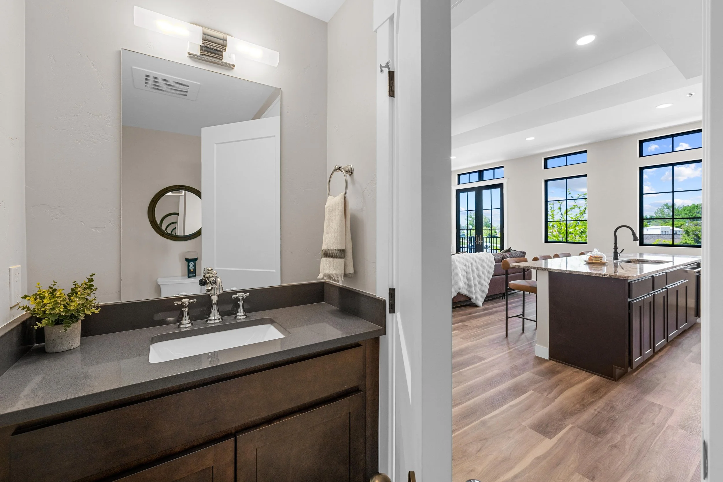 View of a bathroom vanity with a sink, dark wood cabinet, and gray countertop, next to an open doorway leading into a bright, modern living area with large windows and a kitchen island.