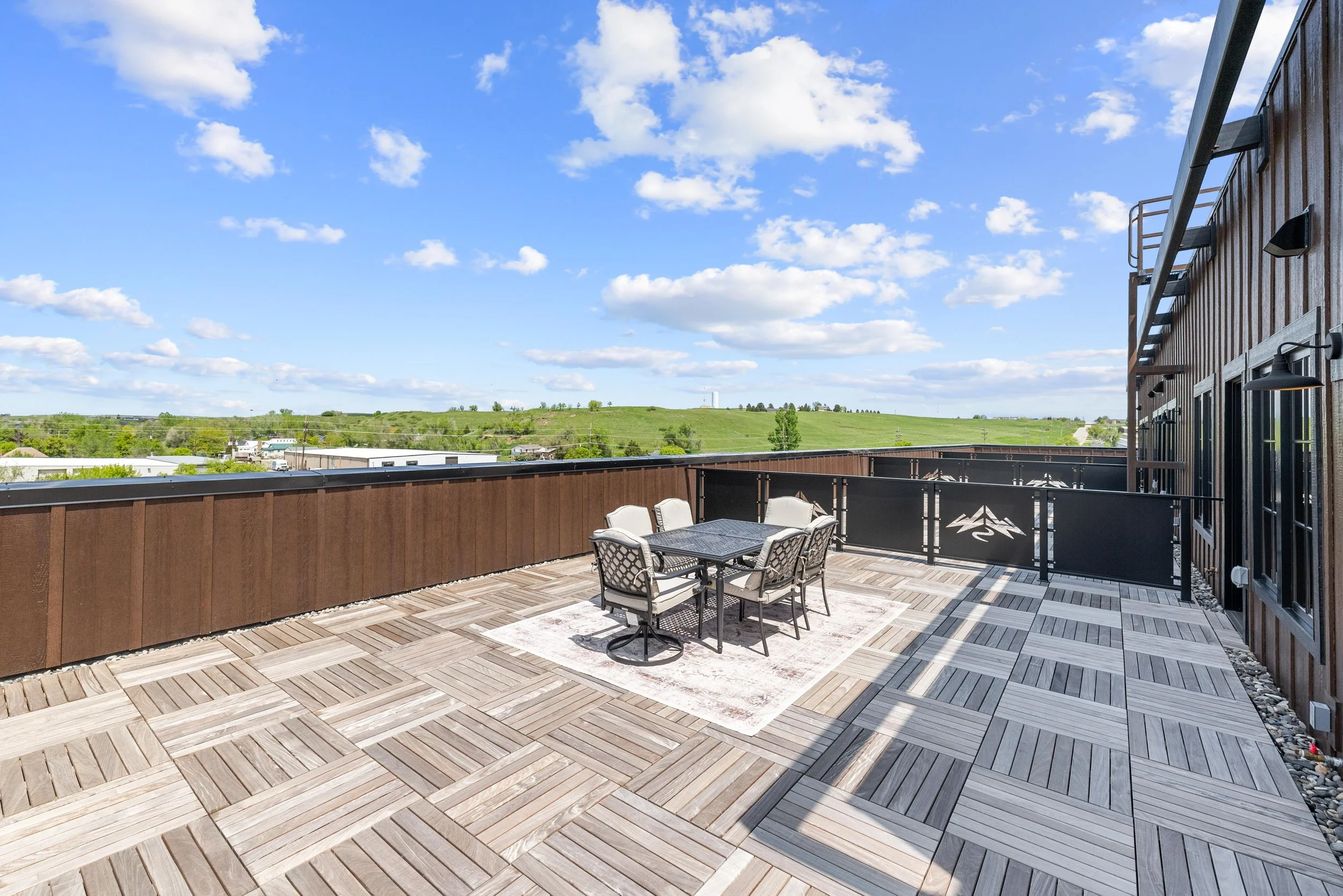 Spacious rooftop terrace with outdoor dining table and six chairs, enclosed by a wooden and metal railing, overlooking green fields under a partly cloudy sky.
