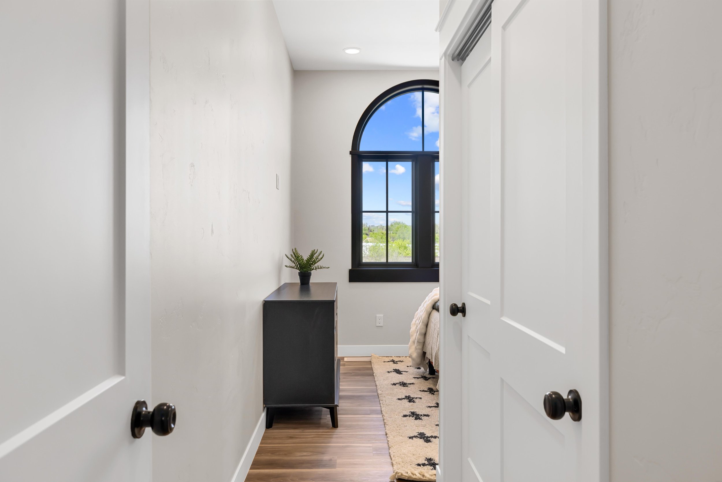 View of a small bedroom with a large arched window showing blue sky and white clouds, white walls, black trim around the window, a gray dresser with a potted plant, and a beige rug with black star patterns, all inside the Ice Haus Condos