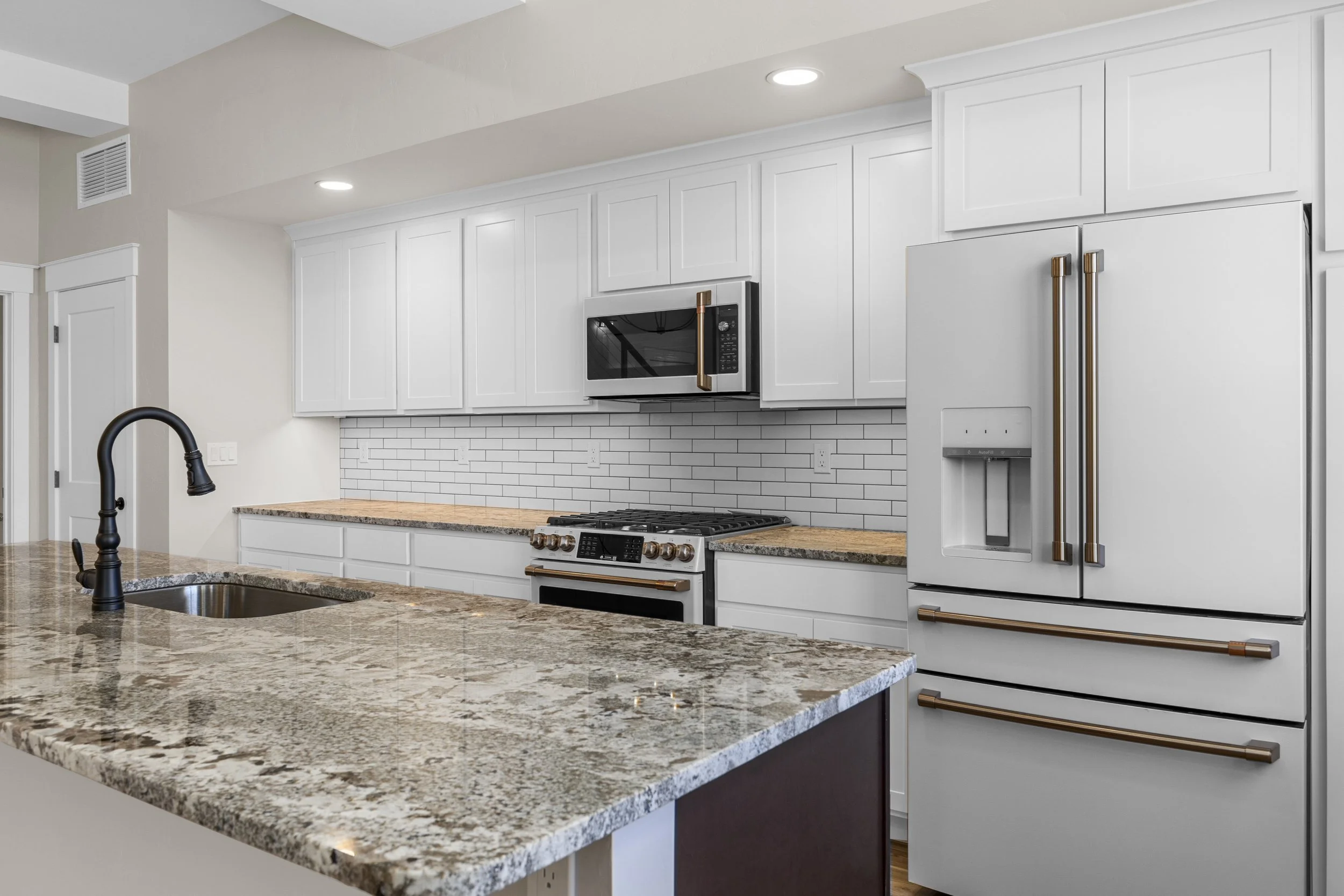 Modern kitchen with white cabinets, granite countertop island with a black faucet, built-in microwave, oven, and large refrigerator with gold handles, white subway tile backsplash, and recessed ceiling lights.
