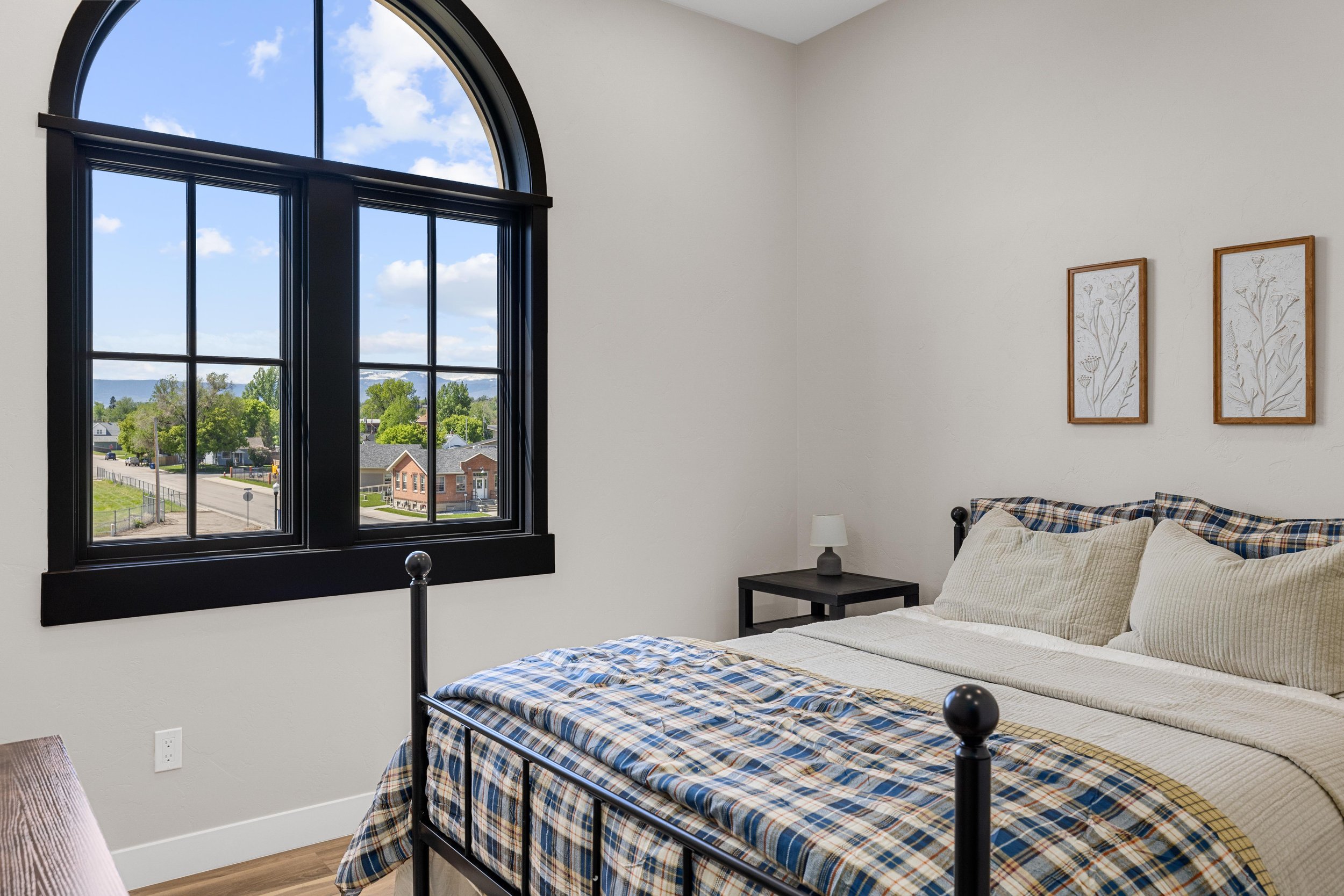 A bedroom with a large arched window, a black-framed window with multiple panes, a black metal bed frame with plaid bedding, two beige pillows, a black nightstand with a small white lamp, and two framed botanical prints on the beige wall.