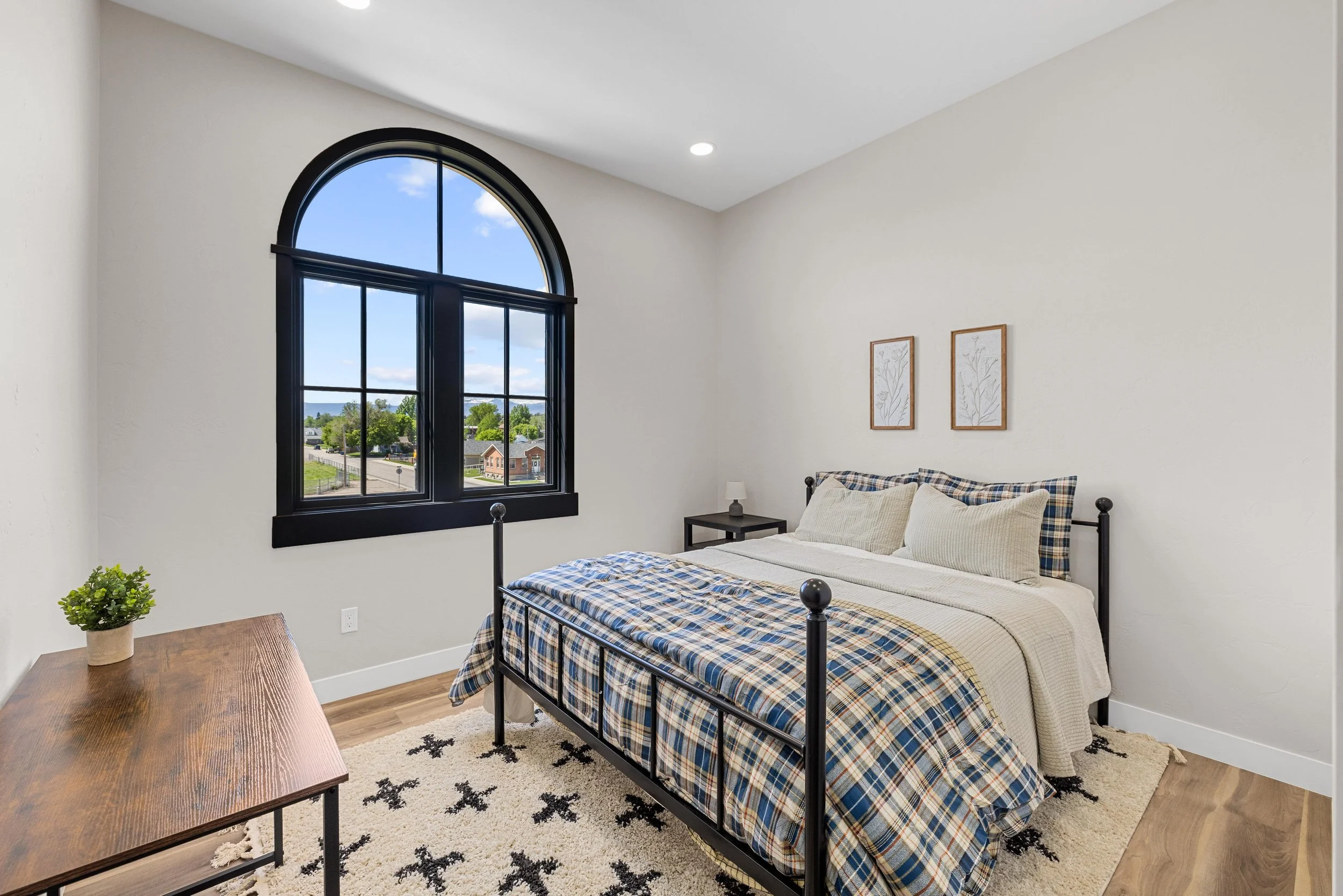 Bedroom with black iron bed frame, cream and plaid bedding, two framed botanical prints on white wall, black window frame showing a view of houses and trees, wooden nightstand with small lamp, wooden table with green plant, black and white patterned 