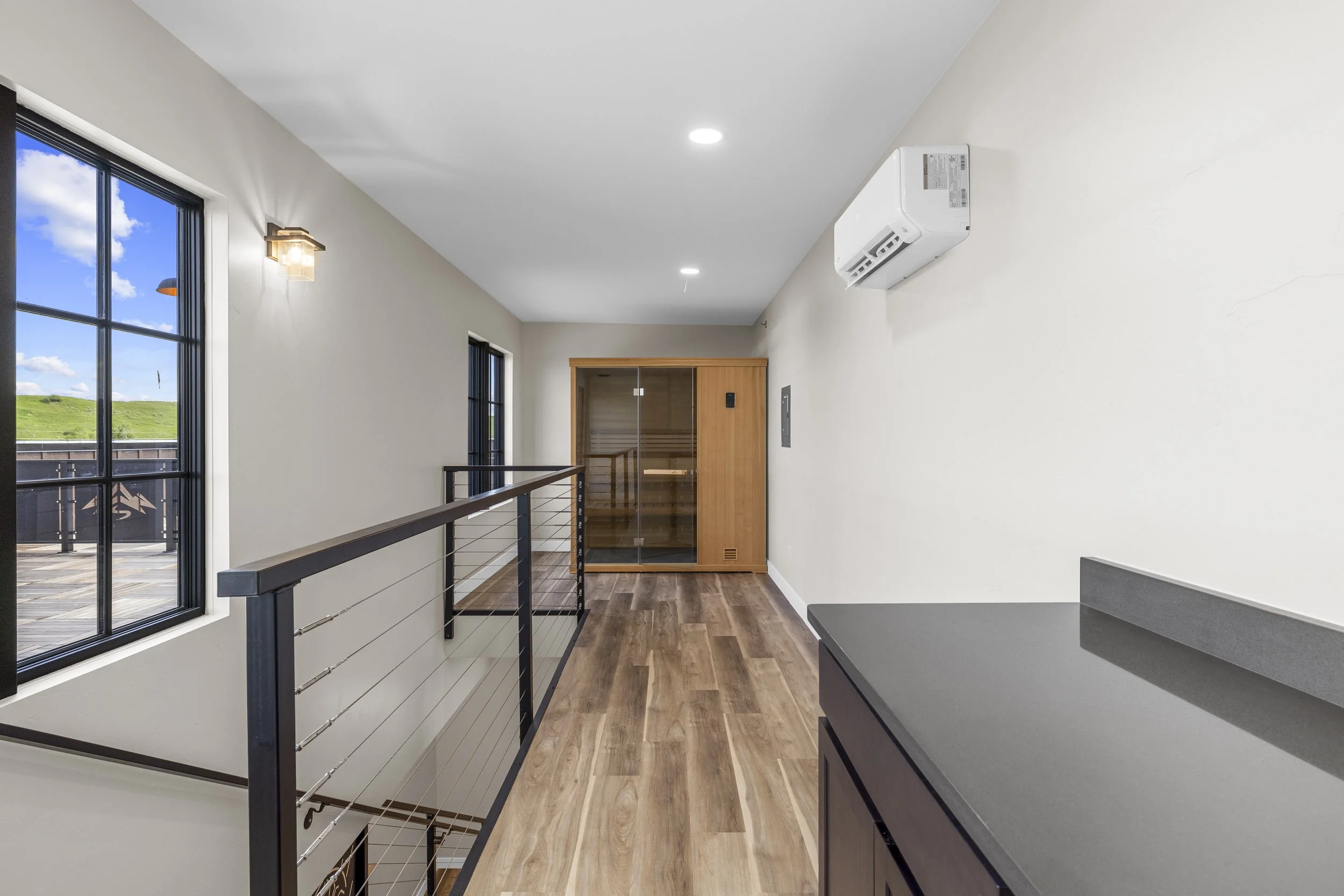 Interior hallway with wooden flooring, windows with black frames, a railing, a sauna with glass doors, and an air conditioning unit on the wall.