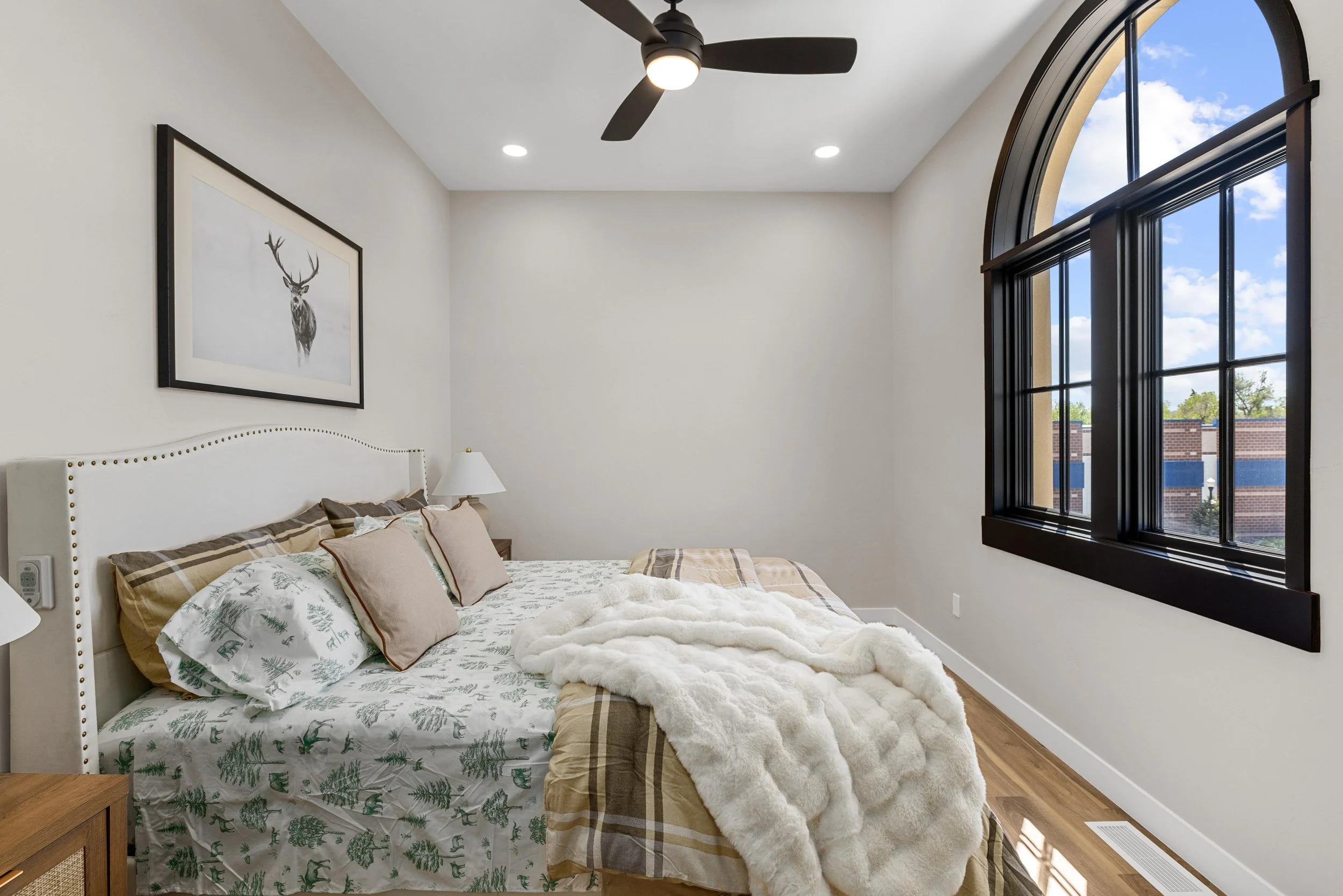 A bedroom with a white bed, beige pillows, a beige and plaid comforter, a fluffy white blanket, a wooden nightstand, a white wall-mounted lamp, a framed picture of a deer, a black ceiling fan, electric outlets, and large arched windows showing a brig