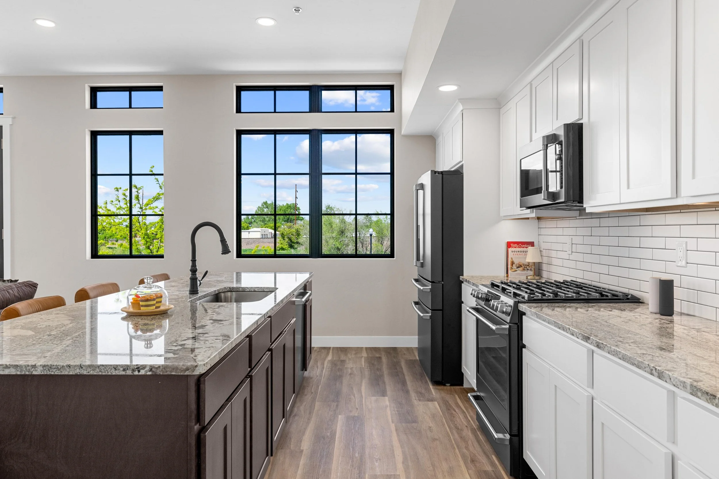 Modern kitchen with white cabinets, granite countertops, black appliances, large windows, and wooden flooring.