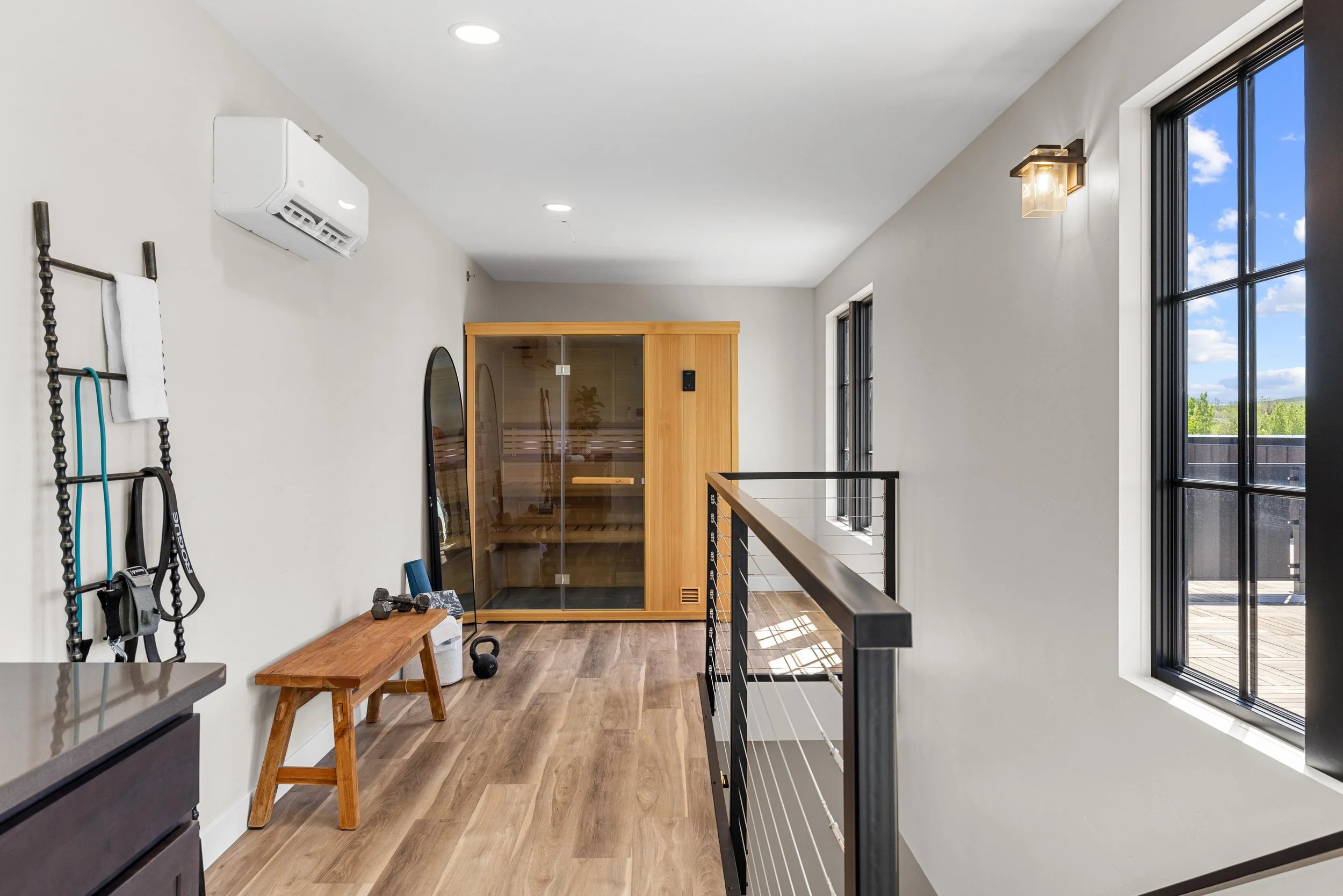 Indoor space with wooden floor, black metal railing, large windows showing outdoors, sauna, exercise bench, kettlebell, dumbbells, and sauna accessories.