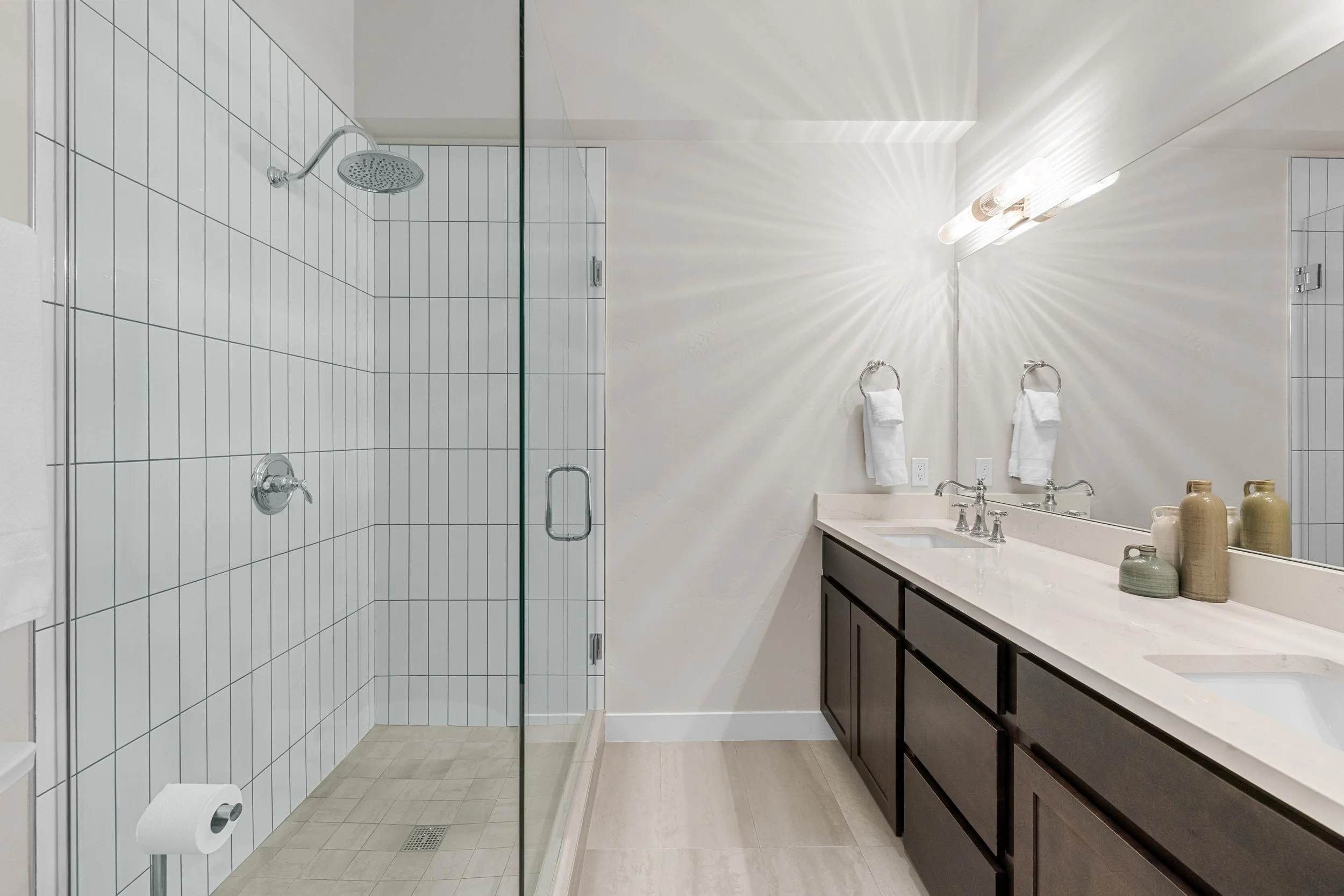 Modern bathroom with a glass-enclosed shower, double sink vanity with dark cabinets, mirror, wall-mounted light fixture, and decorative vases on the countertop.