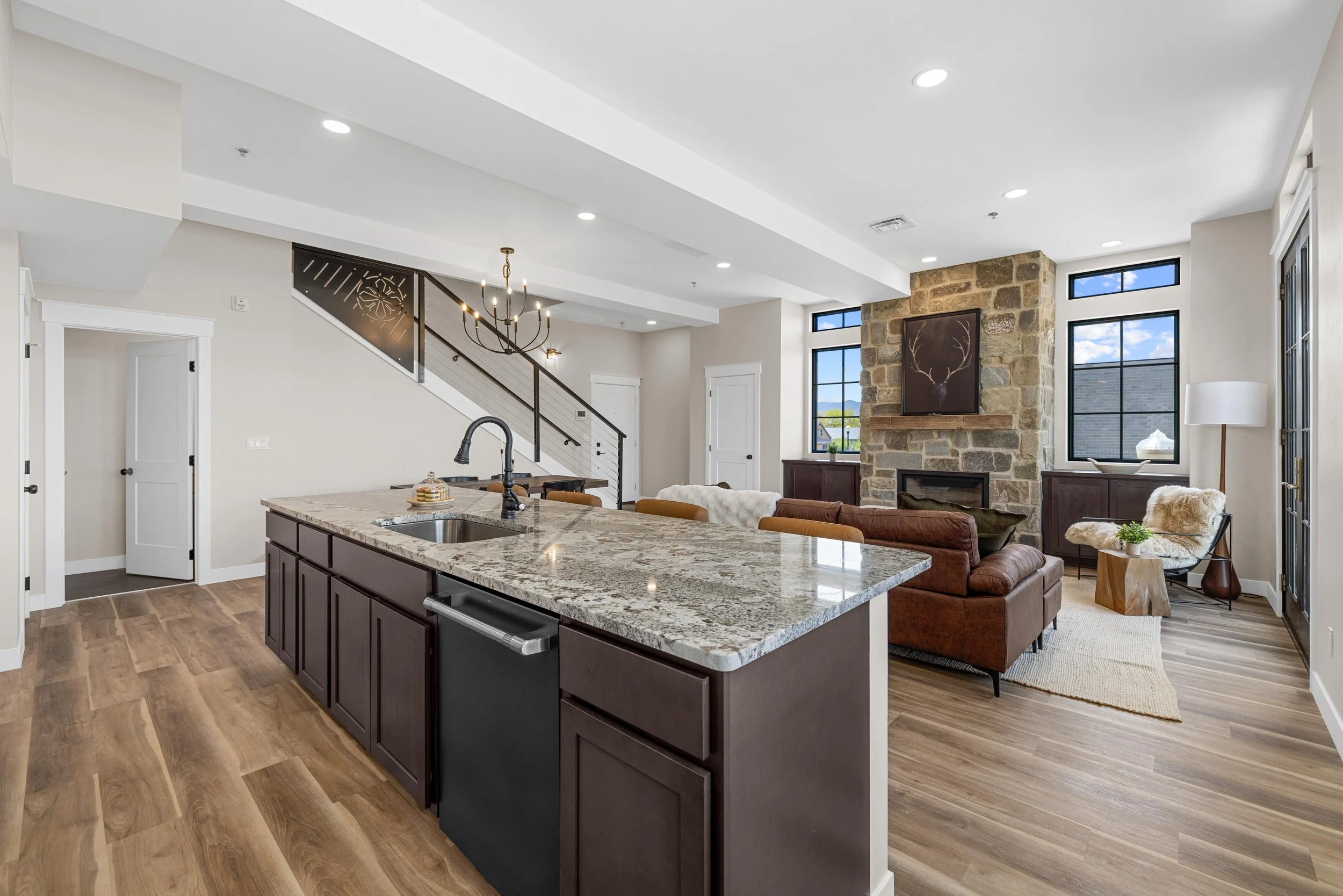 Open-concept living room and kitchen with stone fireplace, large windows, and wooden flooring.