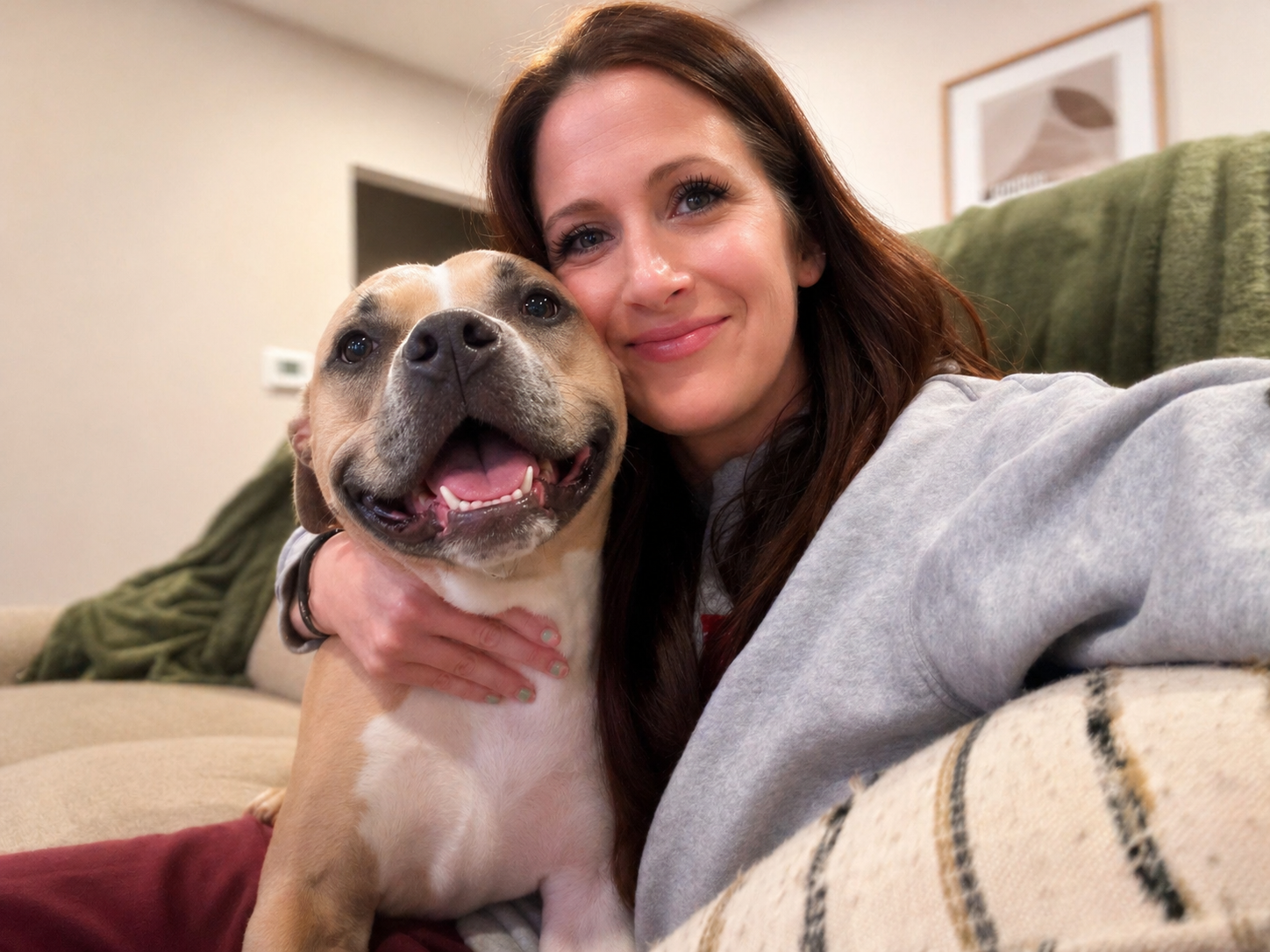 A woman with long brown hair smiling, hugging a happy, tan and white dog on a beige couch in a cozy living room.