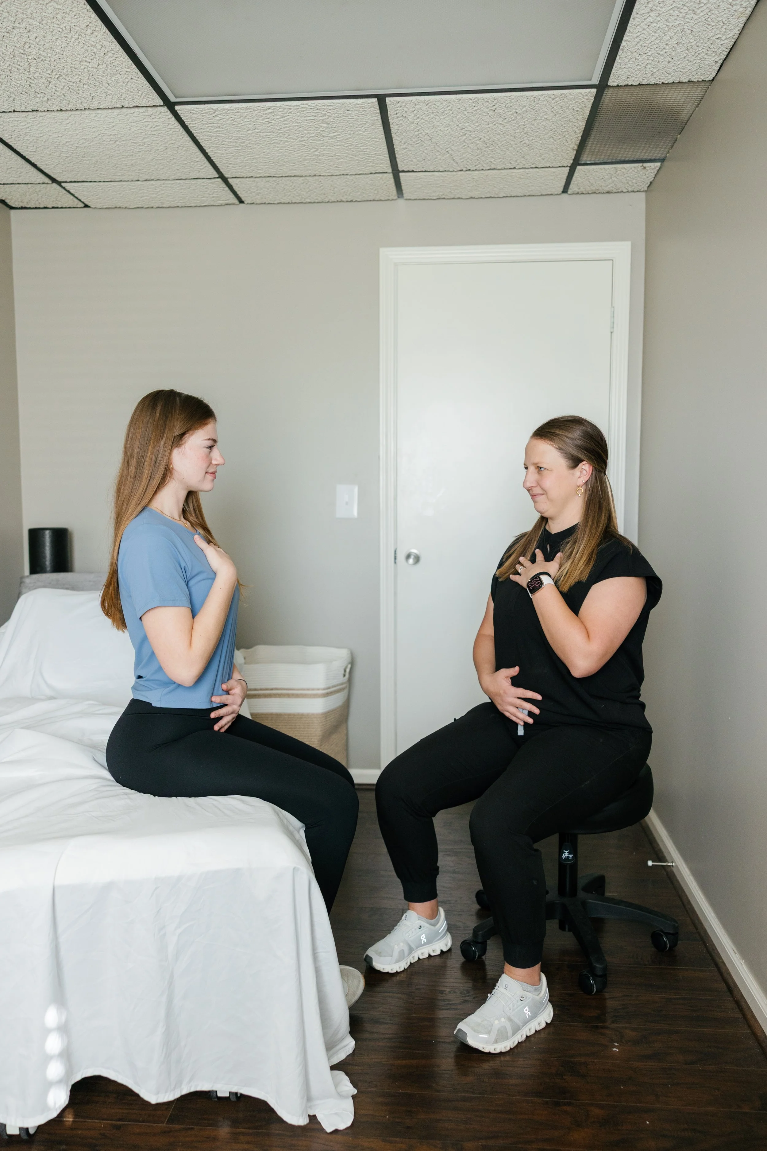 Two women are sitting facing each other with their right hands over their chests, practicing or demonstrating deep breathing exercises in a small, relaxed room with a white door, a bed with a white cover, and a basket.