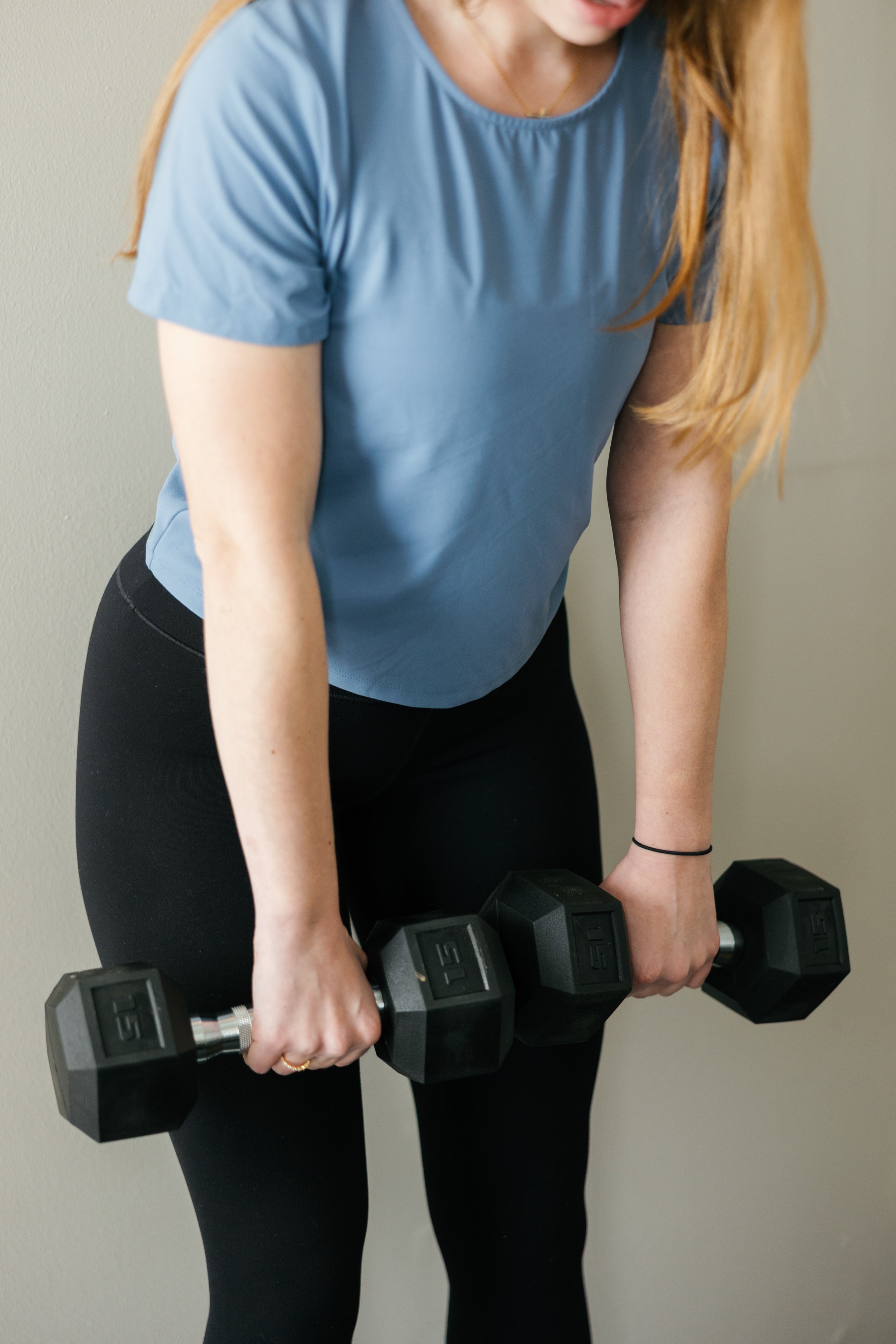 Woman in a blue shirt and black pants lifting black dumbbells with silver handles.