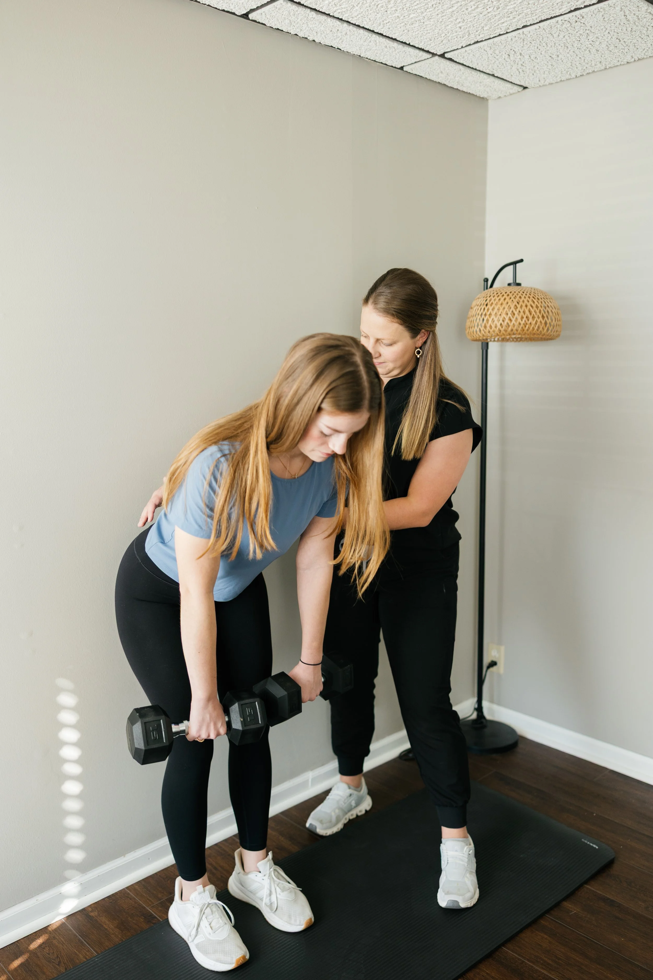 Two women are doing a workout with dumbbells in a gym. One woman is bent over lifting dumbbells, the other is standing close by offering support. They are standing on a black exercise mat.