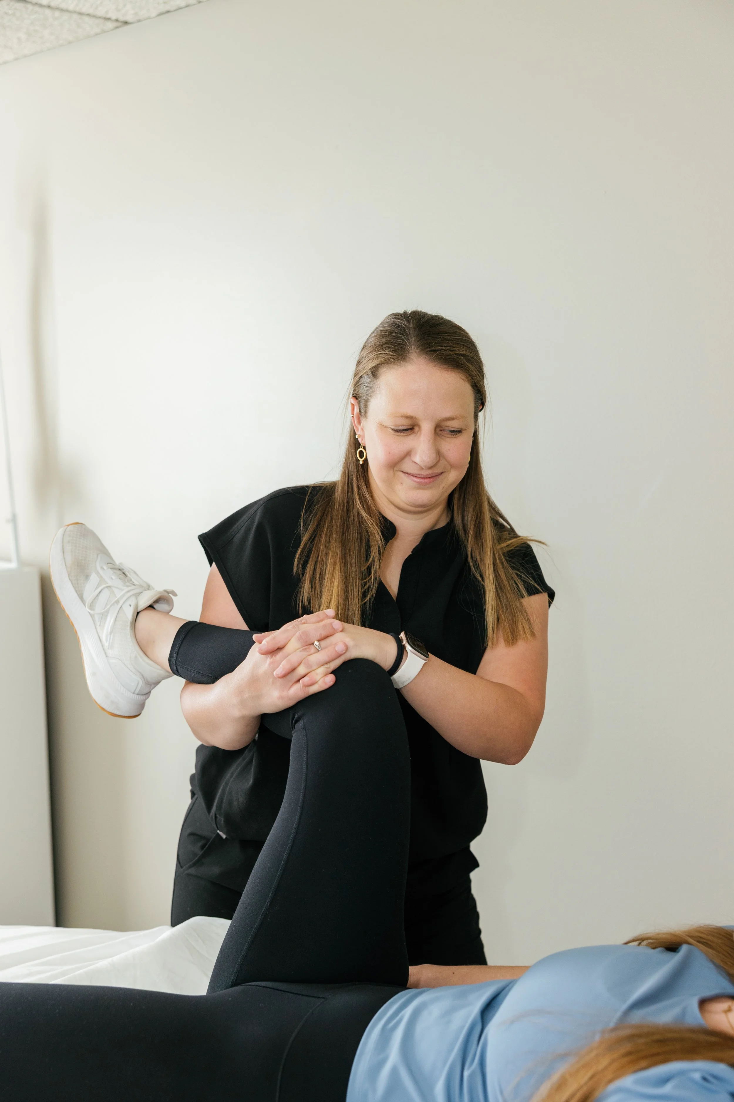 A woman physical therapist is working on a patient's hip in a physical therapy setting. The person is lying on a treatment table, with their foot resting on the woman's arm.