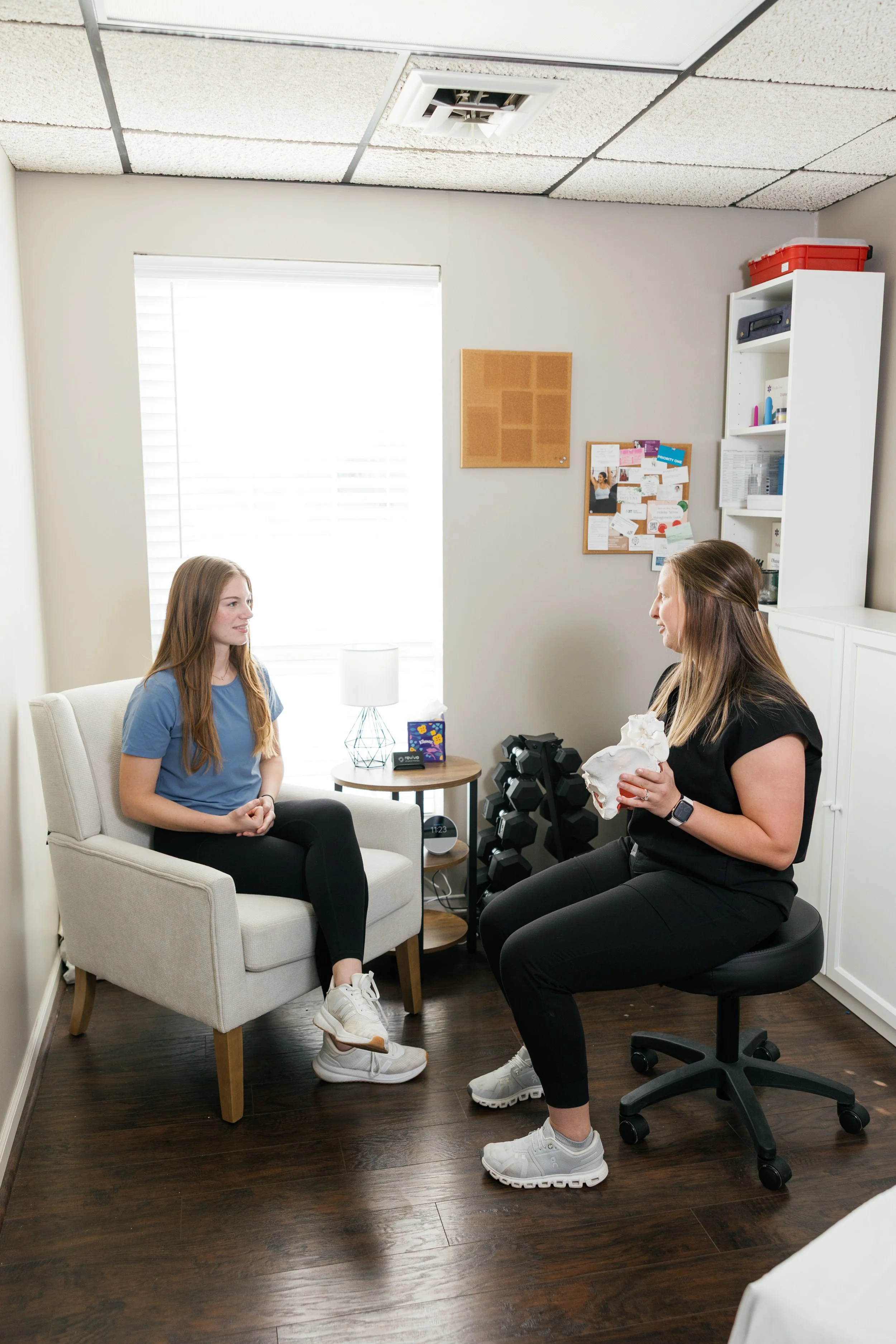 Two women having a conversation in a small office space. One woman with long blonde hair is sitting on an armchair, and the other with long light brown hair is sitting on a rolling office chair, holding a crumpled piece of paper. The office has a small side table, a bookshelf, a corkboard with notices, and a window with blinds.