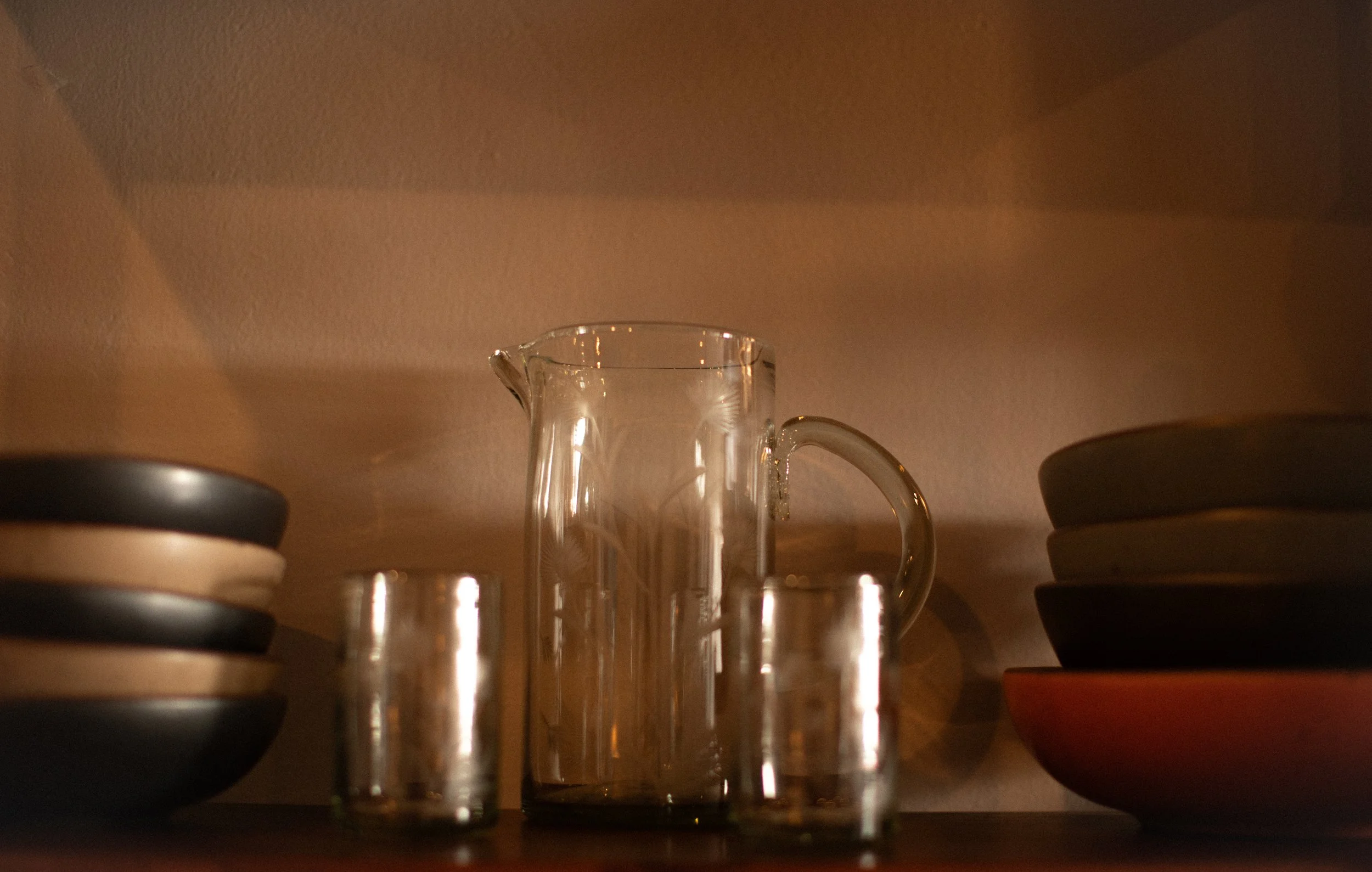 A glass pitcher and four small clear glasses in front of a beige wall, with black and beige plates on the left and stacked dark and red bowls on the right.
