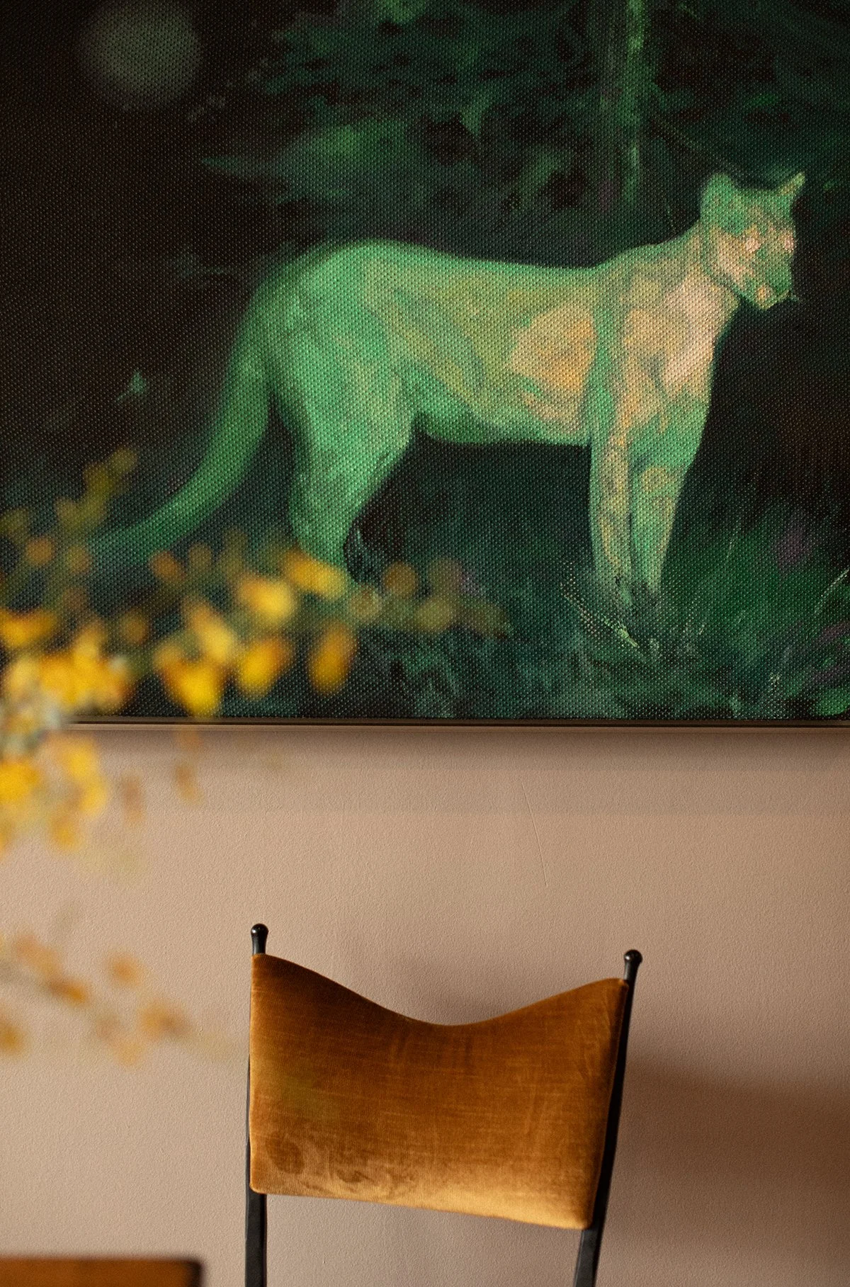 Part of a room showing a wall with a framed picture of a green panther and a chair with a tan cushion in front of it.