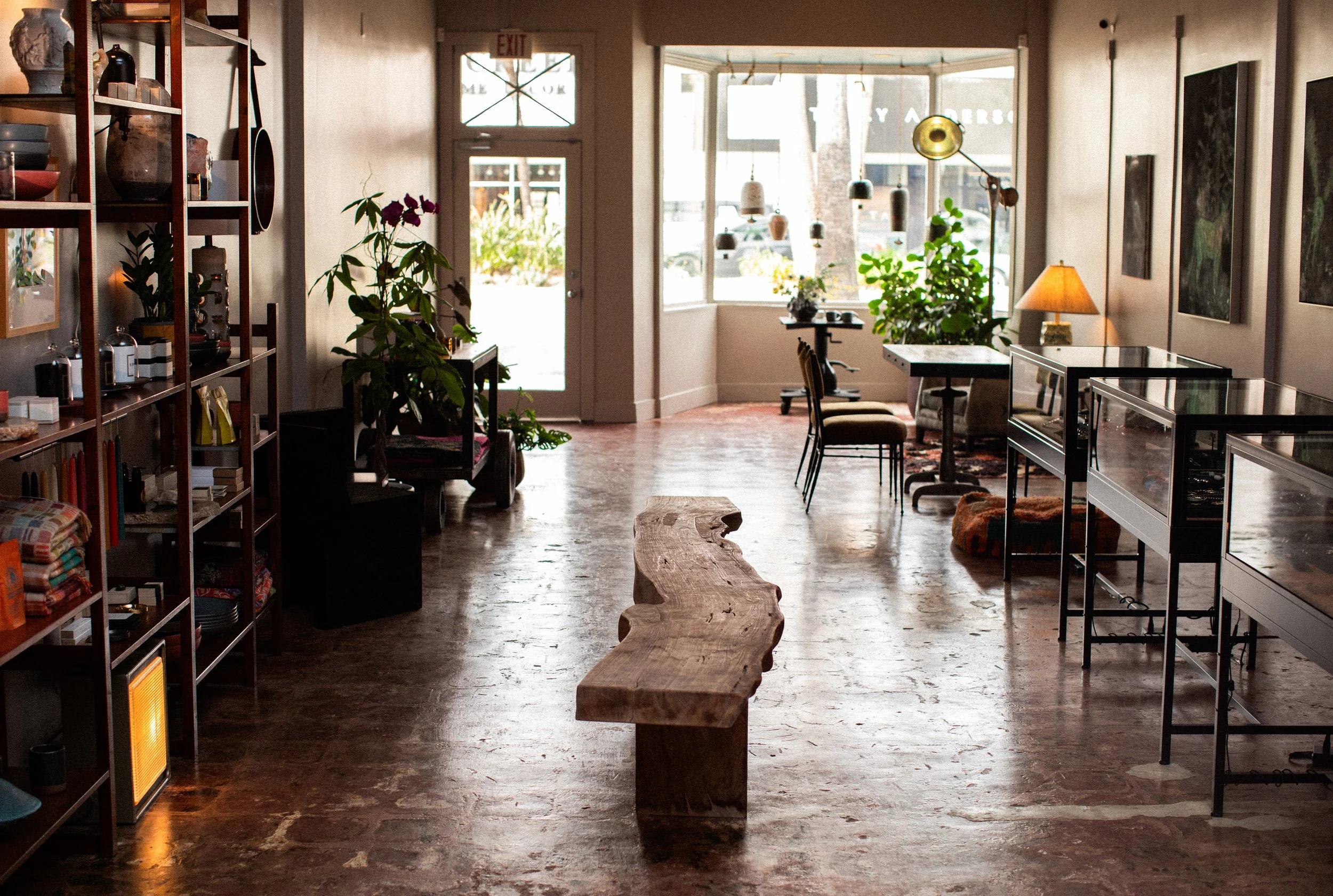 Interior of a cozy shop with concrete flooring, a rustic wooden bench in the center, large windows at the back letting in natural light, plants near the entrance, display cases on the right, and shelves with various items on the left.