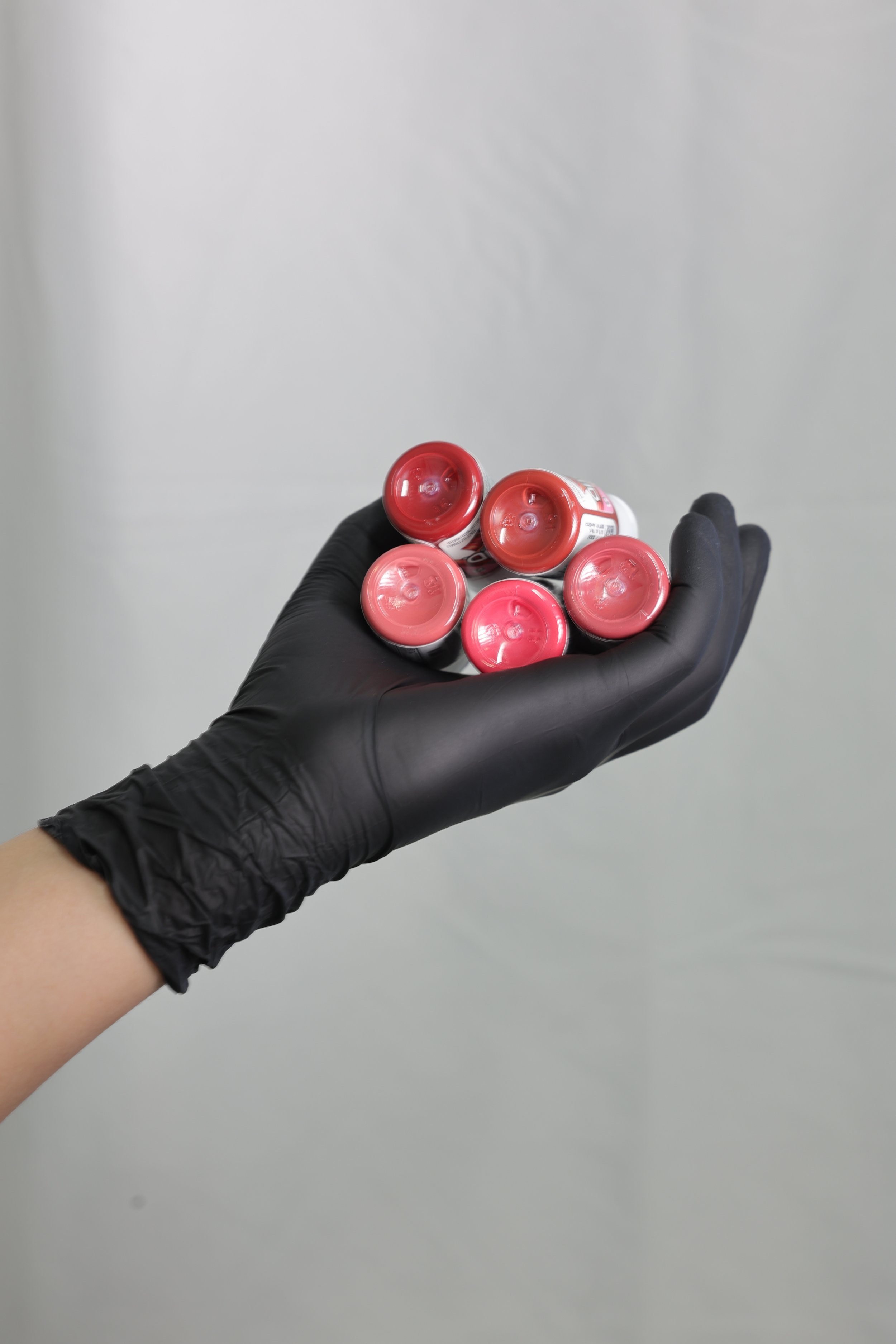 A hand wearing a black glove holding six small containers of pink lip gloss.