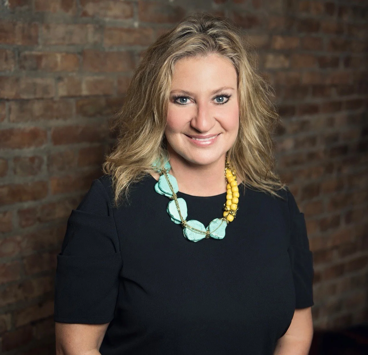 A woman with blonde, curly hair and blue eyes wearing a navy top and a chunky turquoise and yellow beaded necklace, standing in front of a brick wall and smiling at the camera.