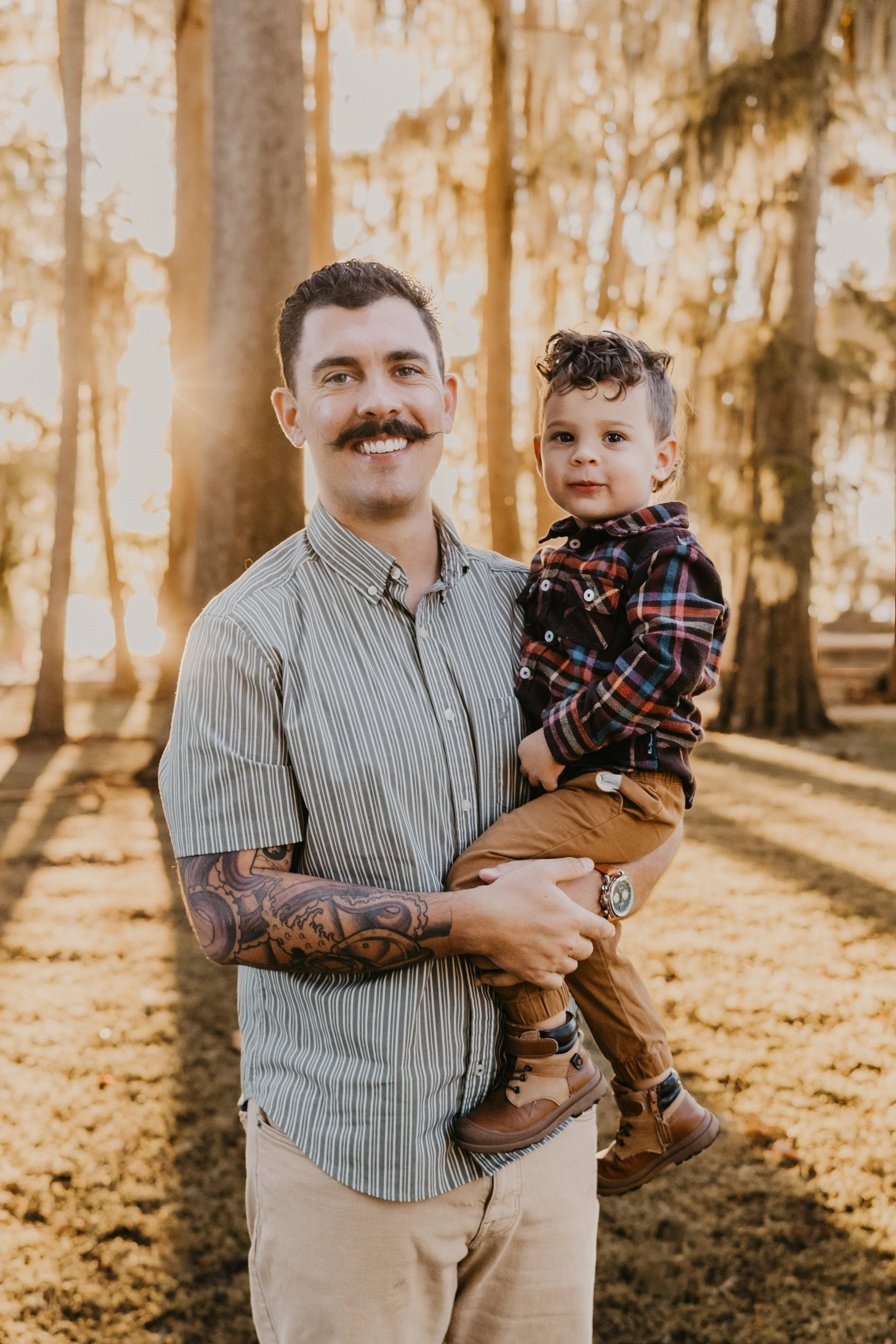 A man with a mustache and a sleeve tattoo holding a young boy in a park with tall trees and golden sunlight filtering through the leaves.