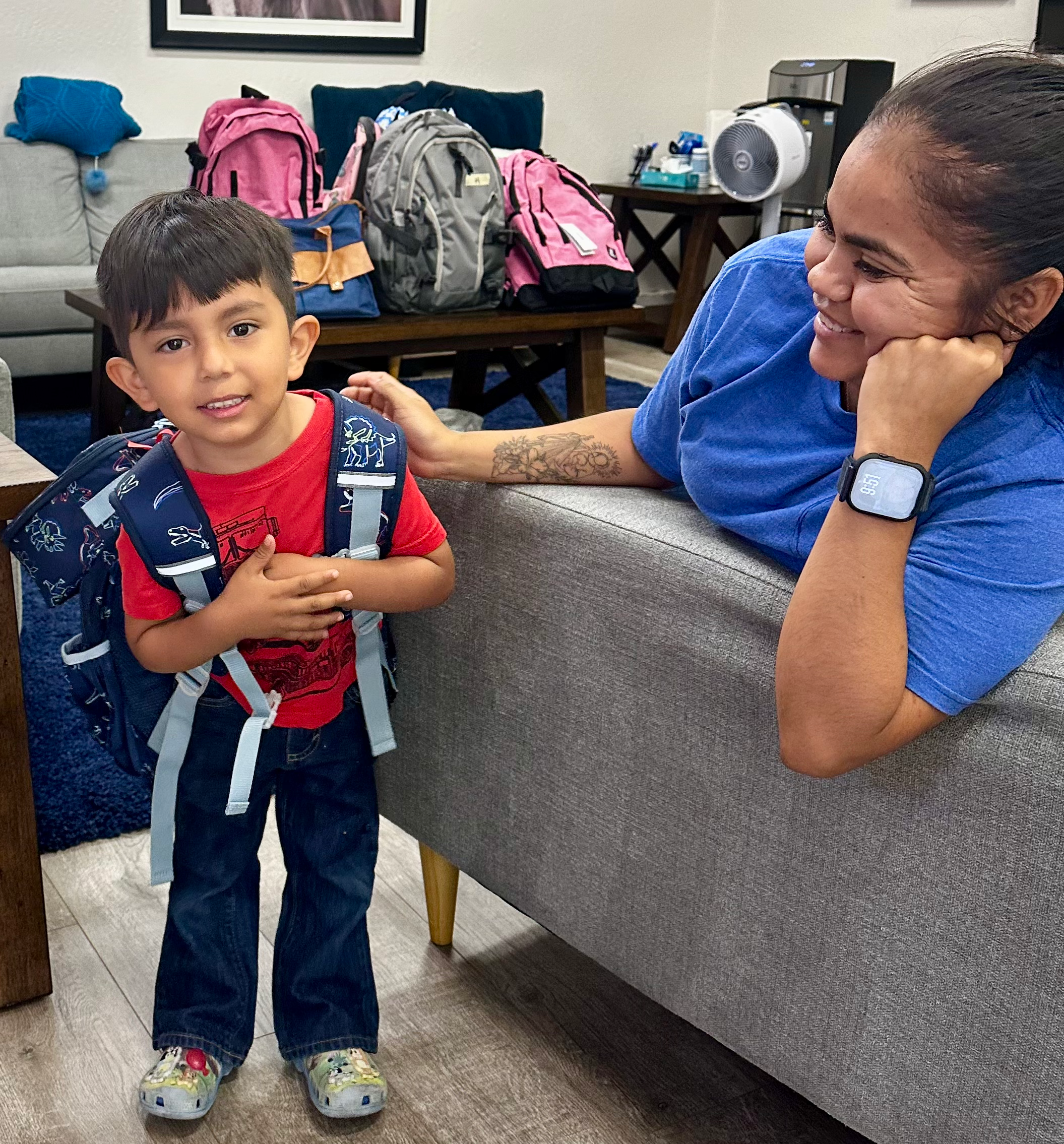 A young boy with a backpack standing next to a woman reclining on a couch, both smiling indoors with backpacks in the background.