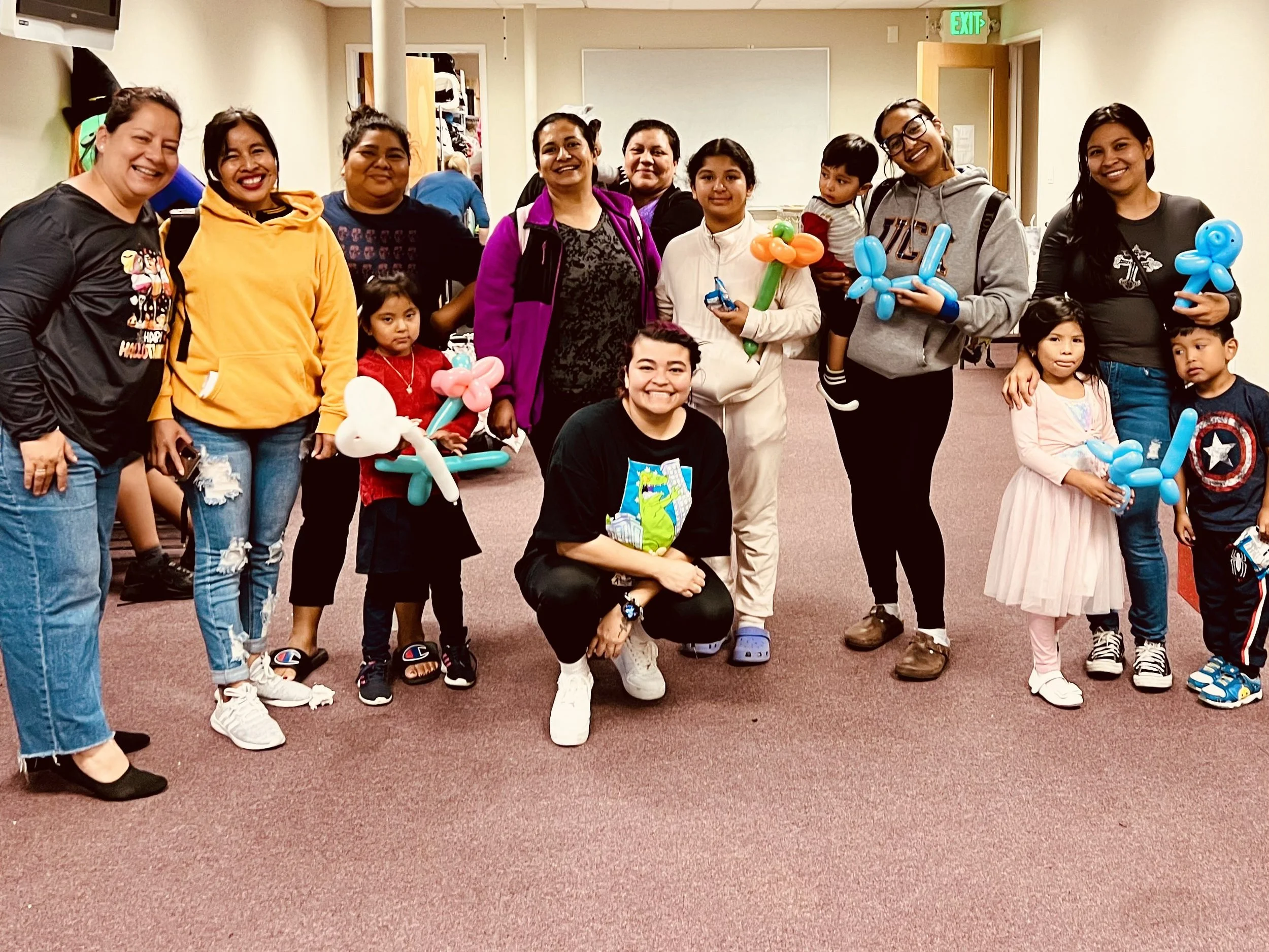 Group of women and children smiling indoors, some holding colorful balloon animals, in a room with beige walls and a red carpet, celebrating or attending a party.