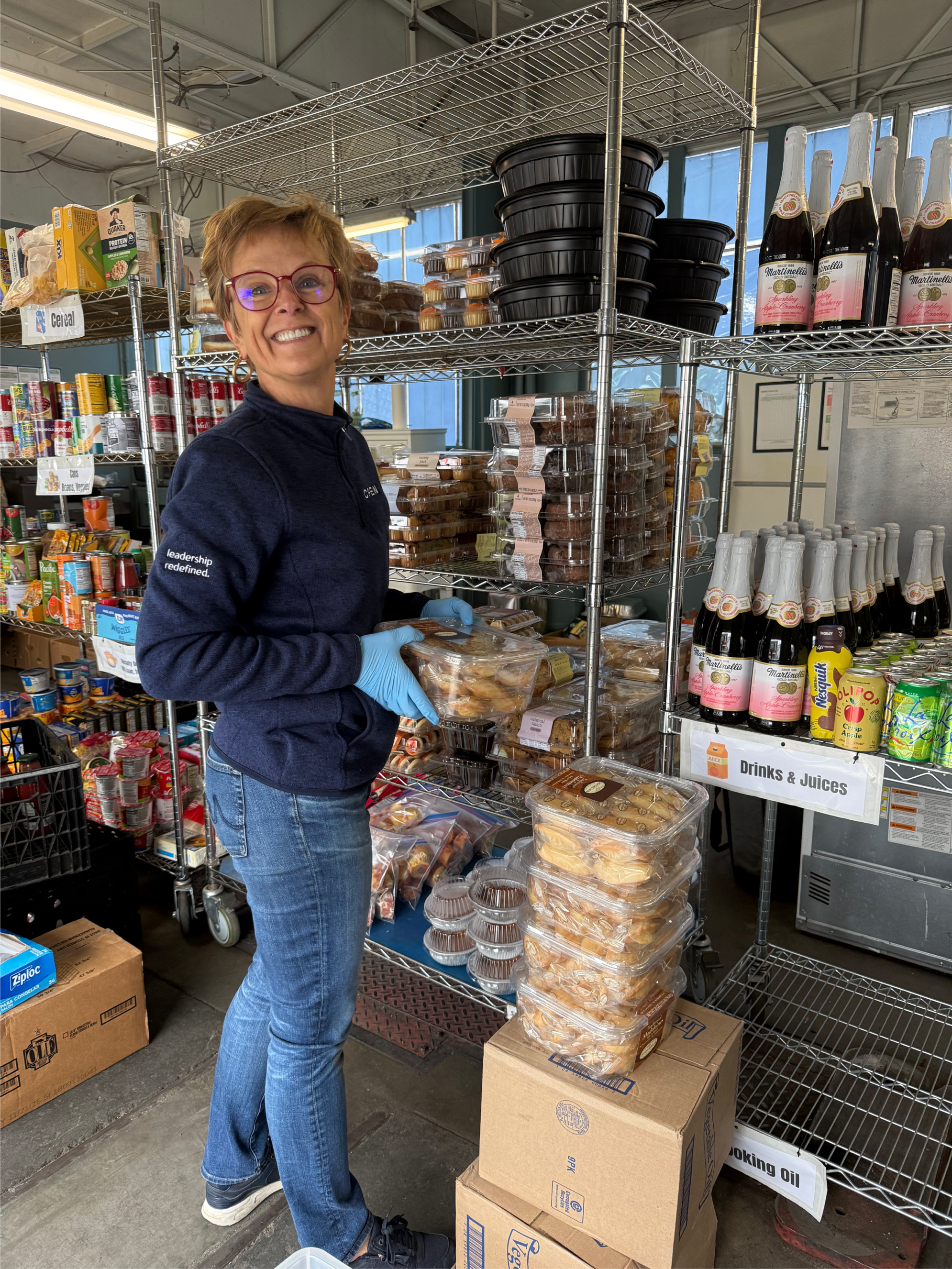 A woman with short hair, glasses, and wearing a dark jacket and blue gloves, stands in a grocery store aisle holding packaged baked goods. Shelves behind her are stocked with baked items, canned goods, and drinks, including bottles of sparkling wine 