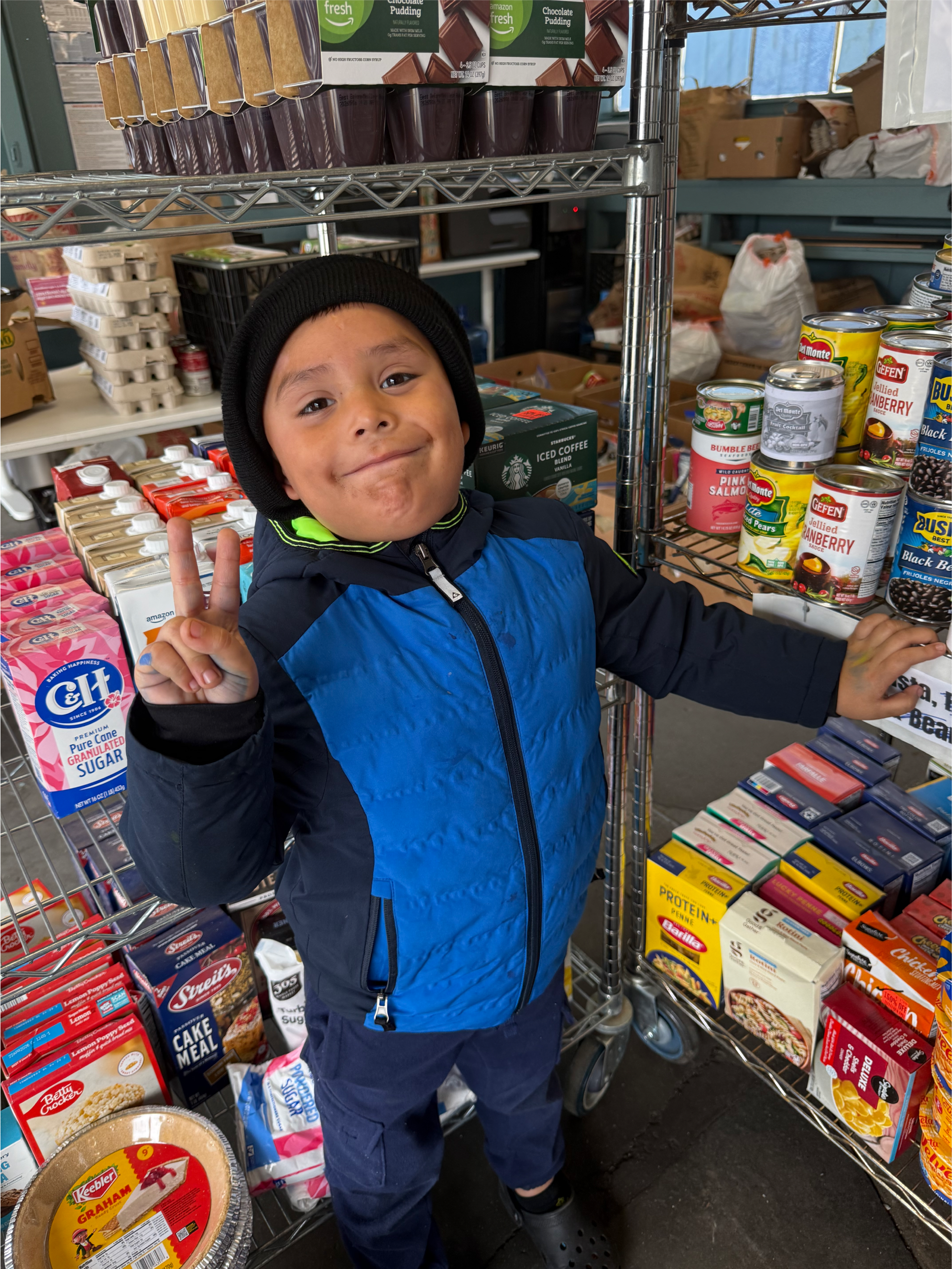 A young boy in a blue and black jacket and black beanie making a peace sign and smiling in a grocery store aisle with canned and boxed food items.