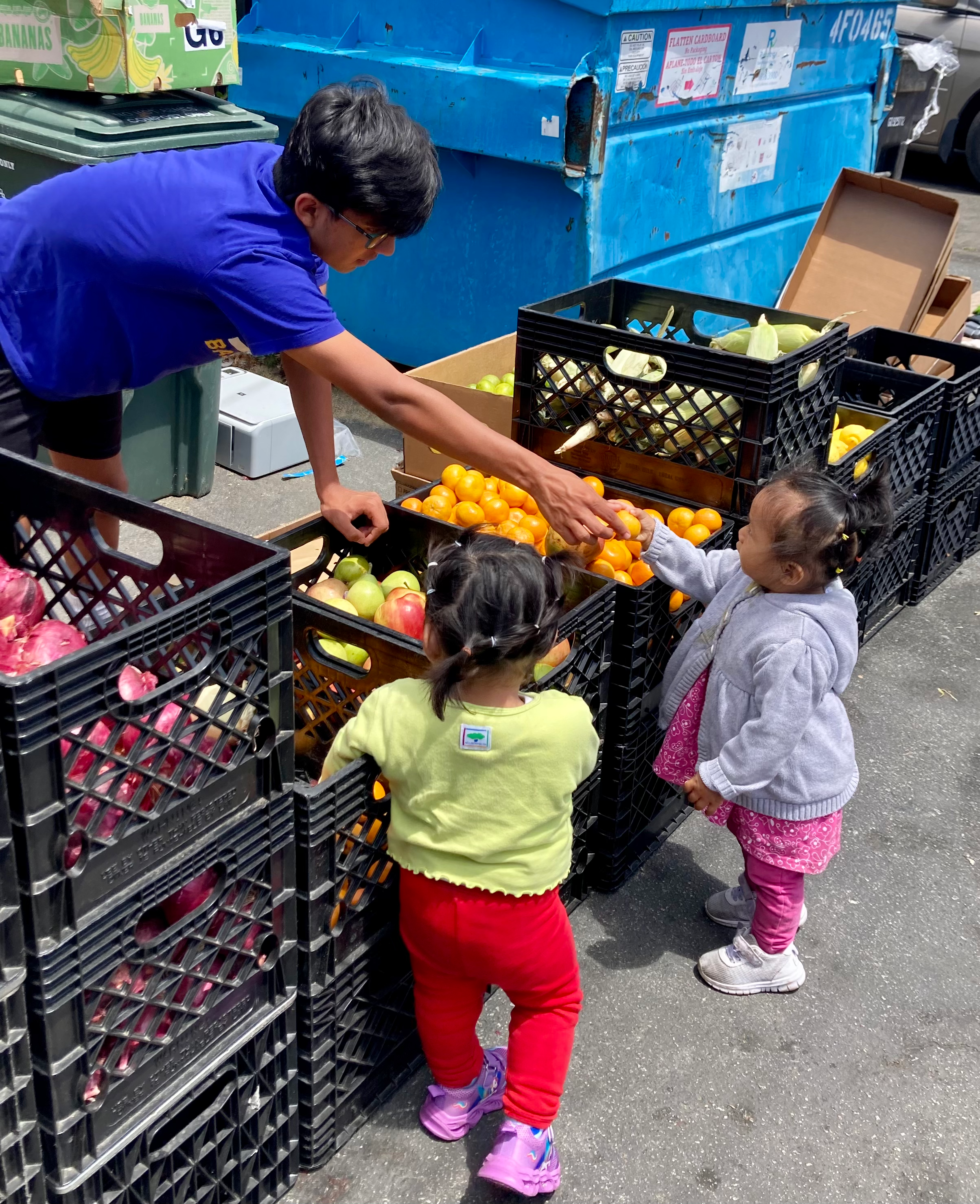 A man is helping two young girls pick oranges at an outdoor produce stand with black crates filled with fruits like apples, peaches, and red onions, beside a blue dumpster.