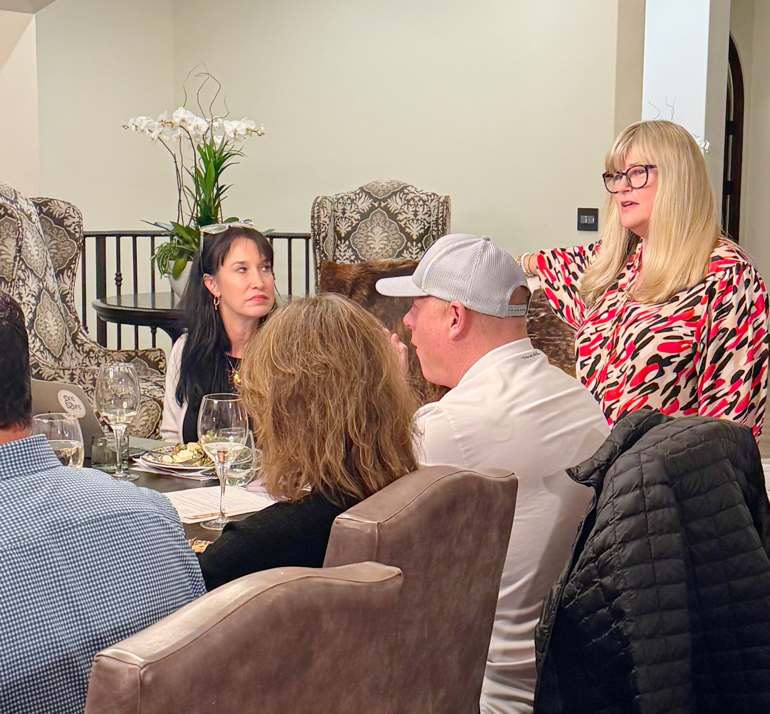 A woman with blonde hair, glasses, and a colorful patterned blouse speaking to a group at a dinner party. A woman with dark hair and earrings listening attentively. Several other guests are seated around a dinner table with wine glasses, plates, and menus.