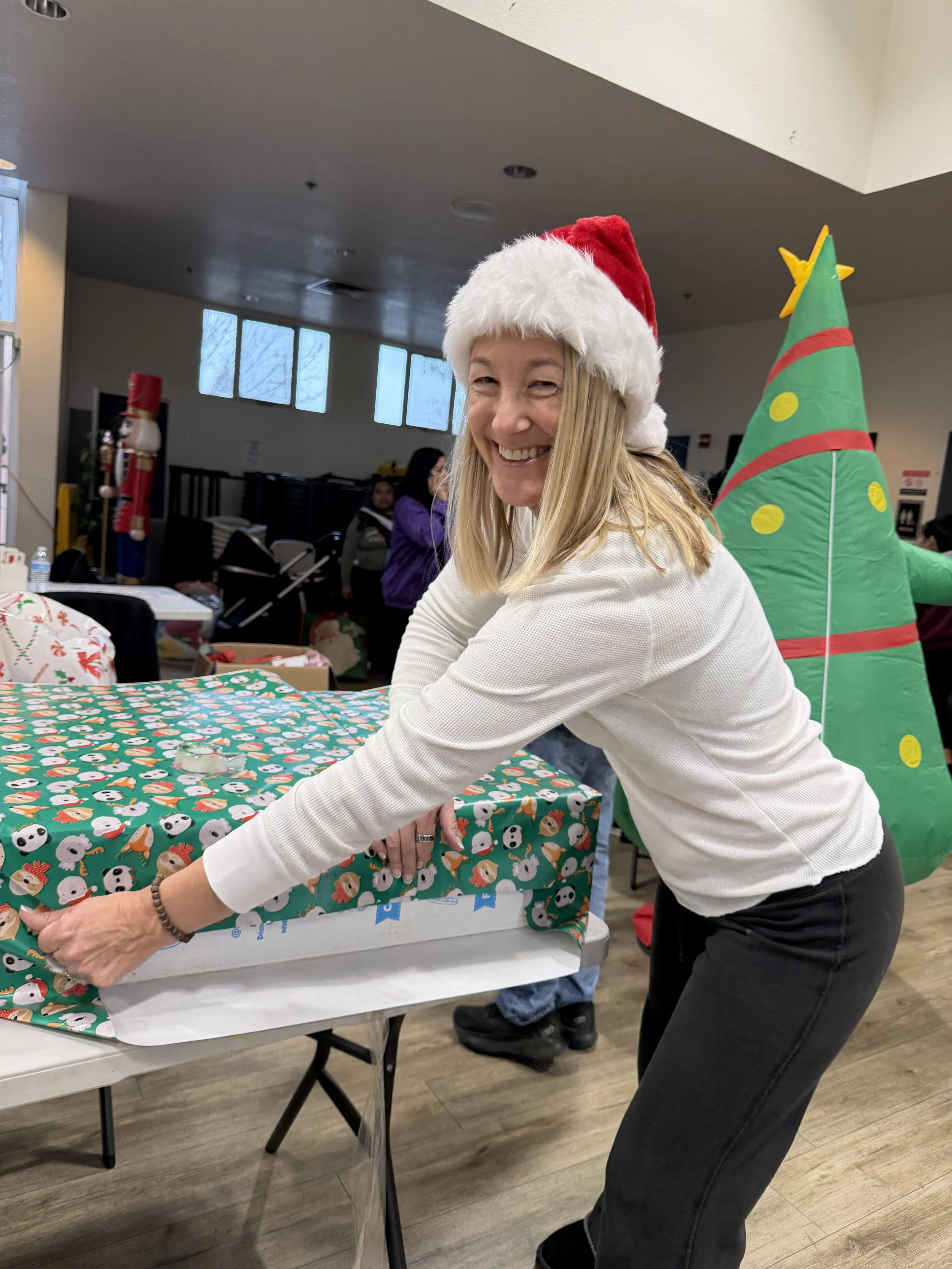 A woman wearing a Santa hat is smiling and wrapping a Christmas gift on a table covered with festive wrapping paper. In the background, there is a decorated Christmas tree, a nutcracker figure, and several people in a room.