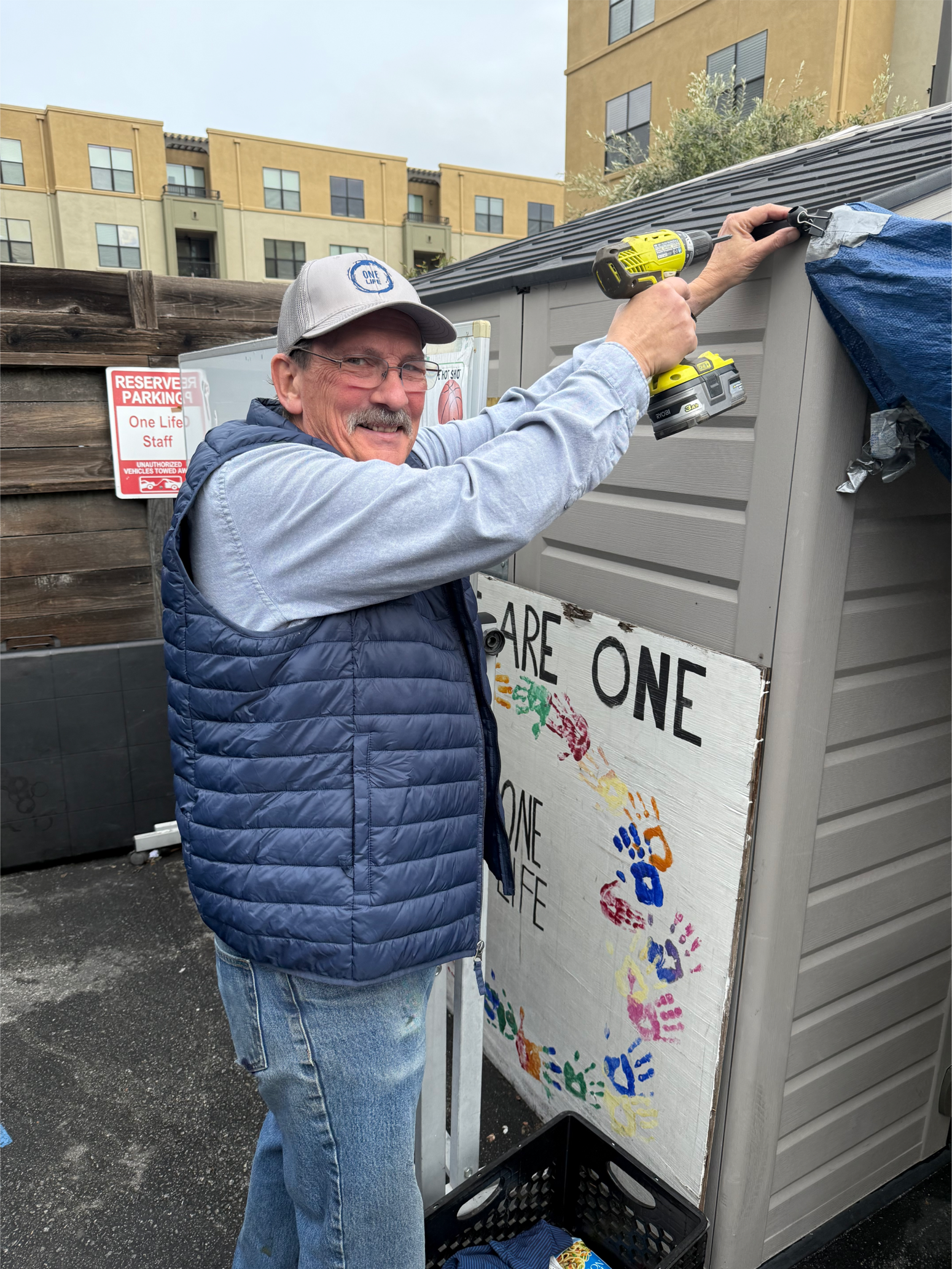 A man with glasses and a gray cap is using a cordless drill to attach a tarp to the side of a shed. He is smiling and wearing a blue puffy vest and a light blue shirt. Behind him are buildings and a sign that reads "Share One" with colorful handprint