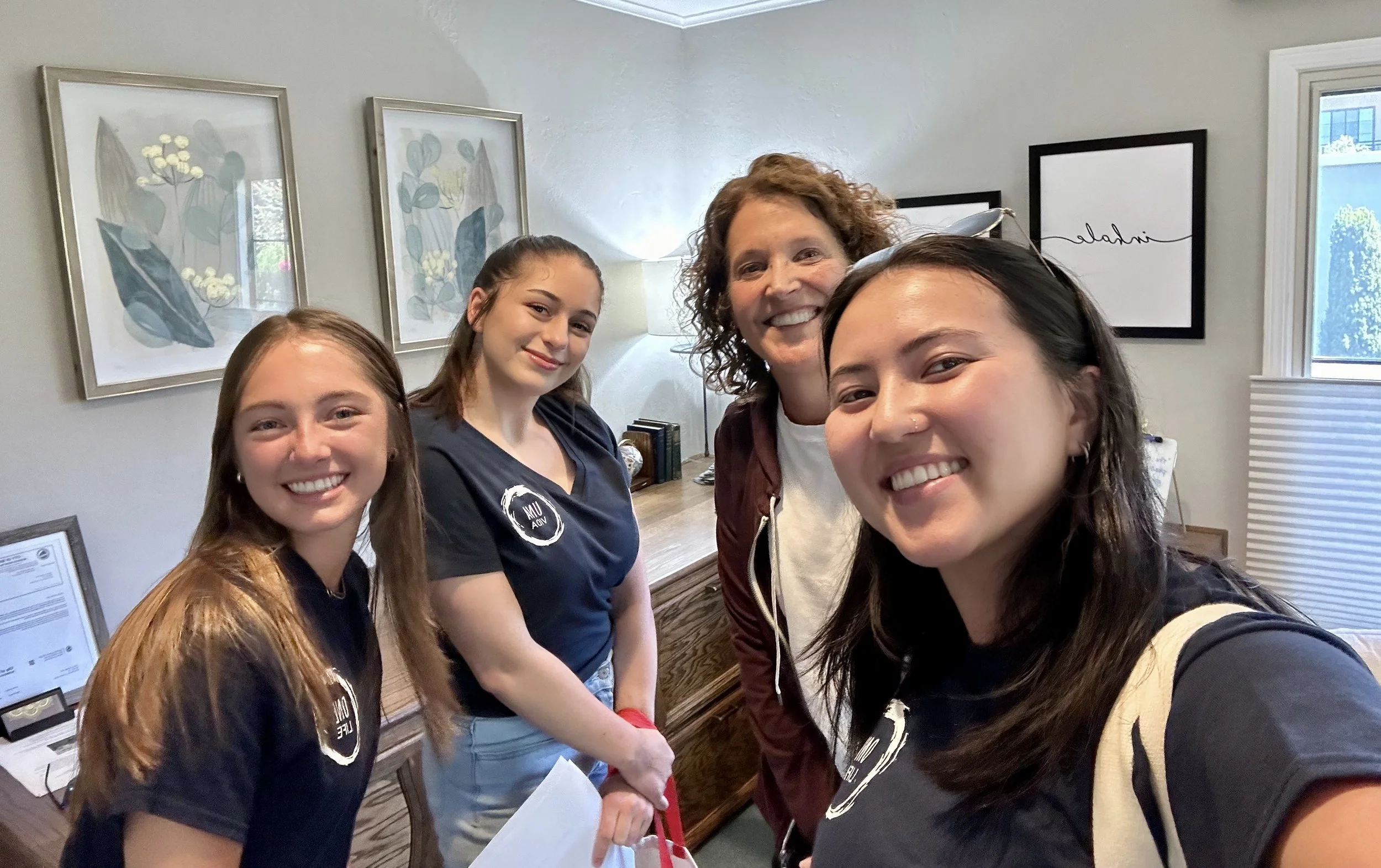 Four women smiling and taking a selfie in an office or home setting with framed artwork and a window in the background.