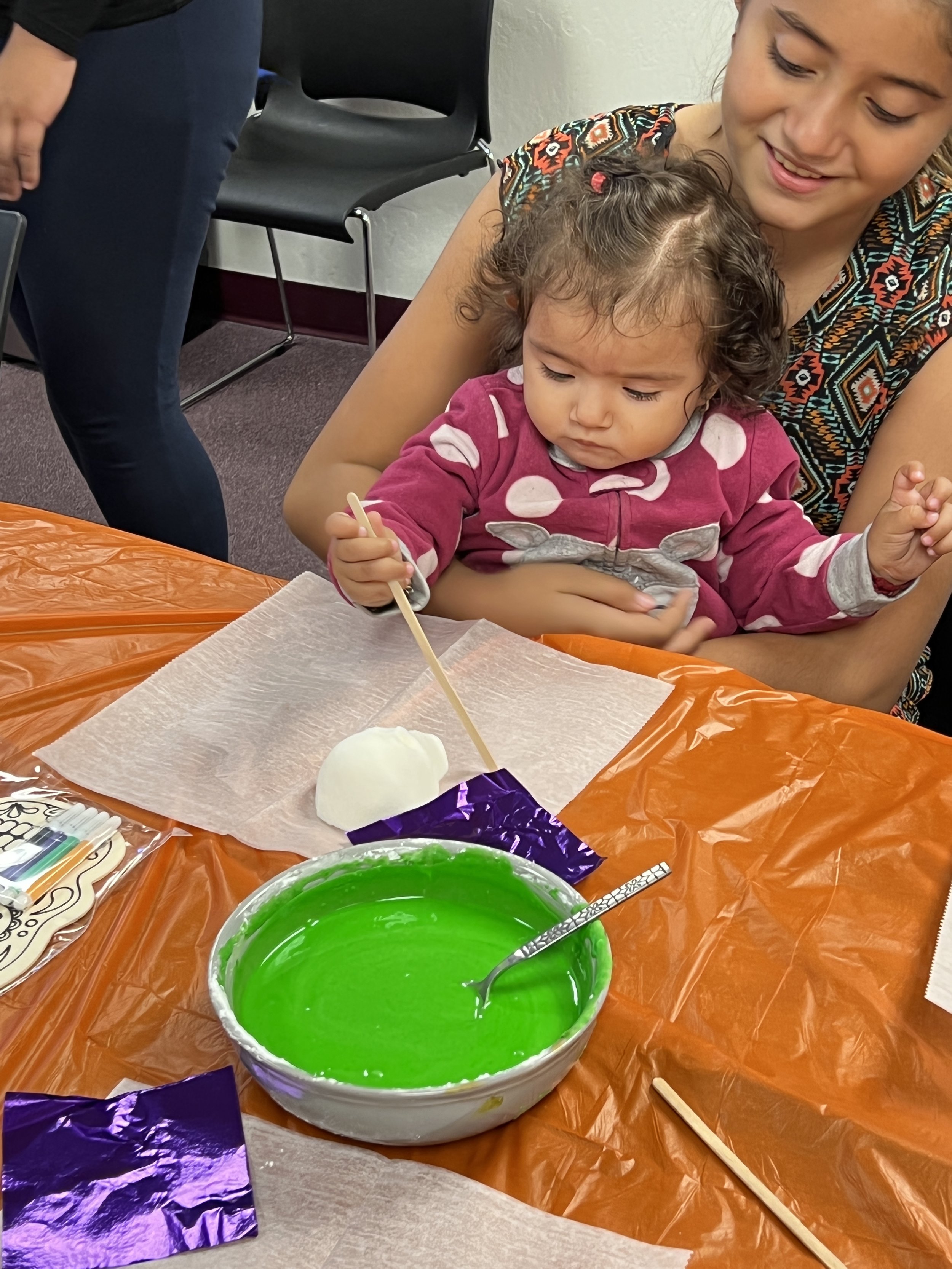 A young girl sitting on a woman's lap, using a stick to poke green slime in a bowl during a sensory activity at a table covered with orange and white paper. The girl is wearing a pink hoodie with polka dots and a gray graphic of a bunny.