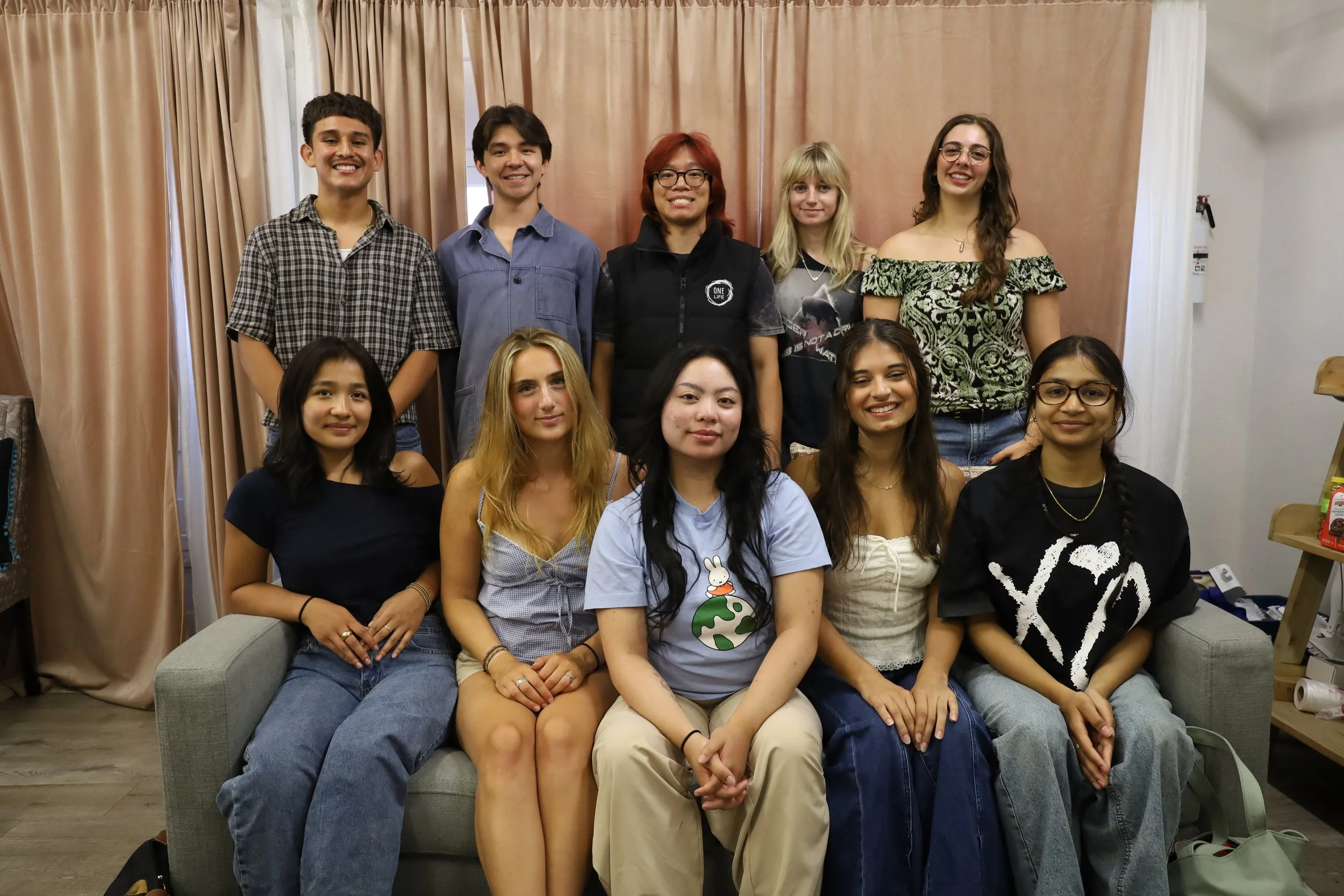 Group of ten young people posing for a photo indoors, some sitting on a sofa and others standing behind, in front of beige and white curtains.