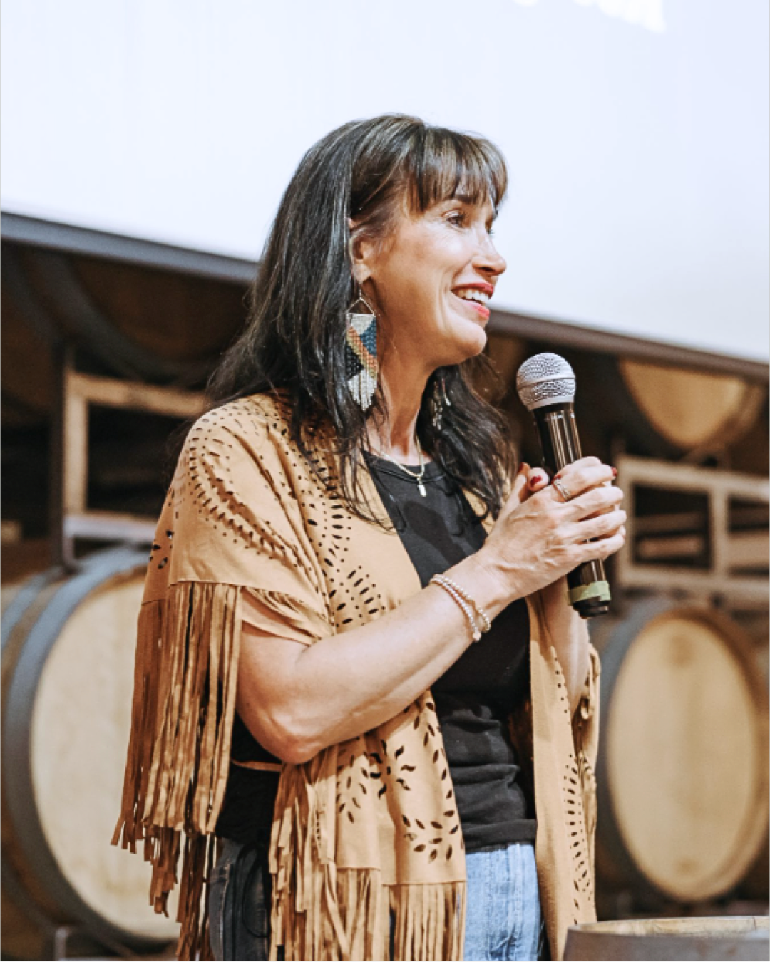 A woman with dark brown hair holding a microphone and speaking in front of a backdrop with large drums.