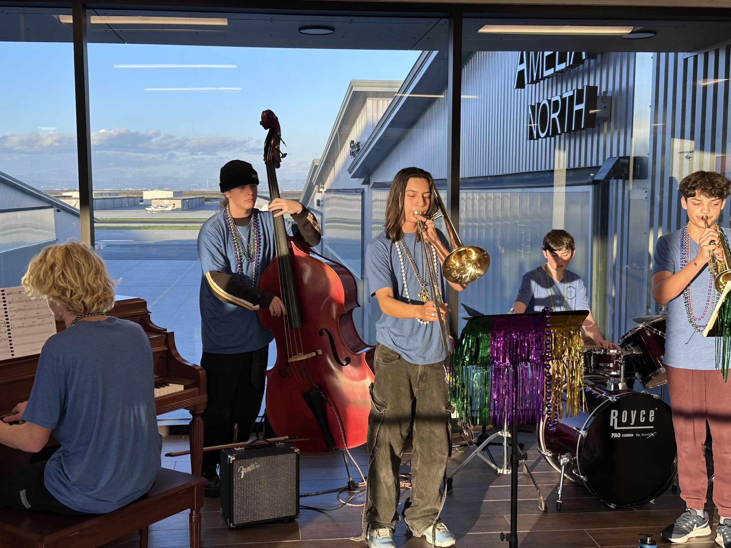 Group of young musicians playing instruments in a room with large windows, an airport runway, and sky visible outside, including a piano, upright bass, trumpet, drums, and a microphone with colorful decorations.