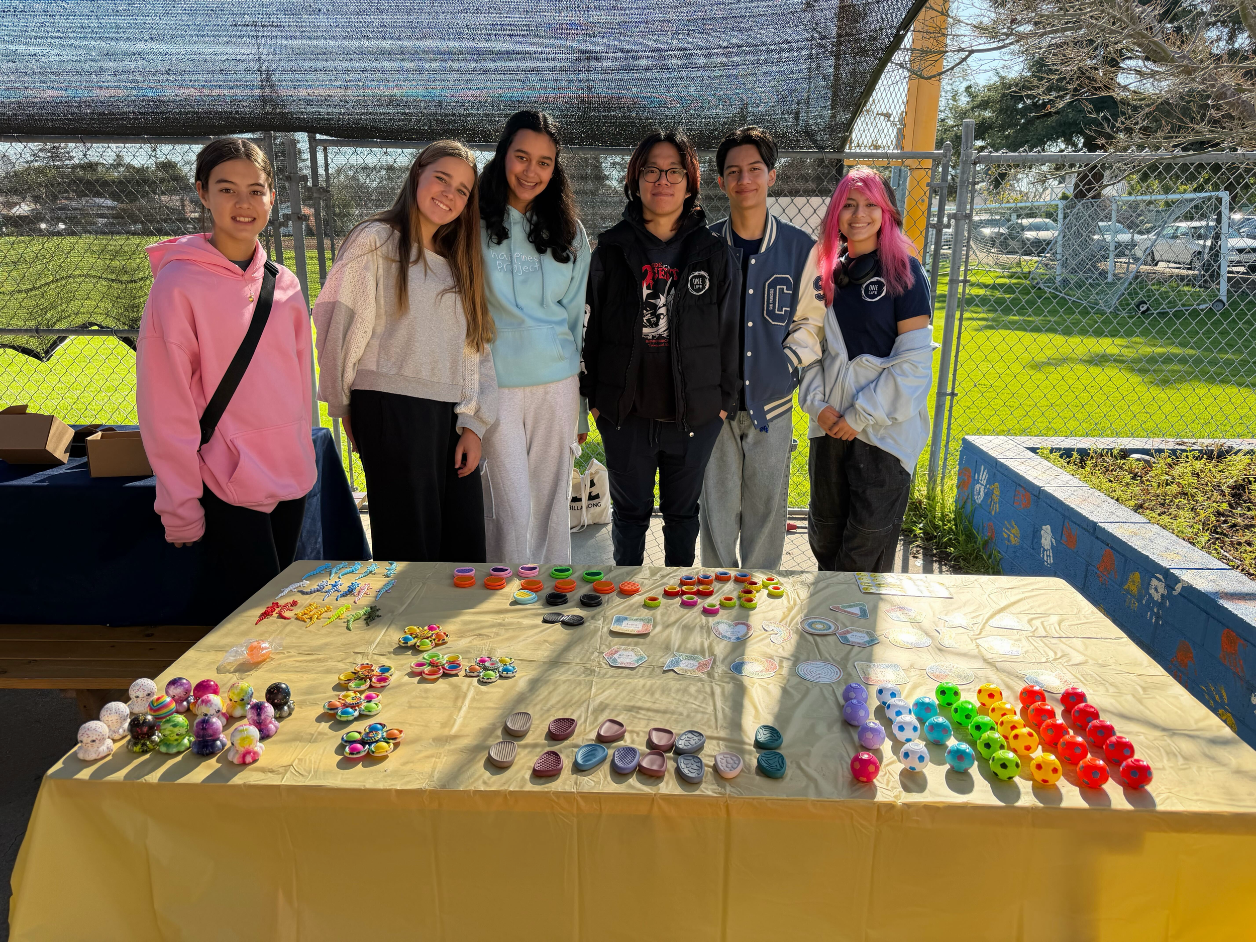 A group of seven young people standing behind a table with various colorful items, outdoors at a fair or event, with a chain-link fence and grassy area in the background.