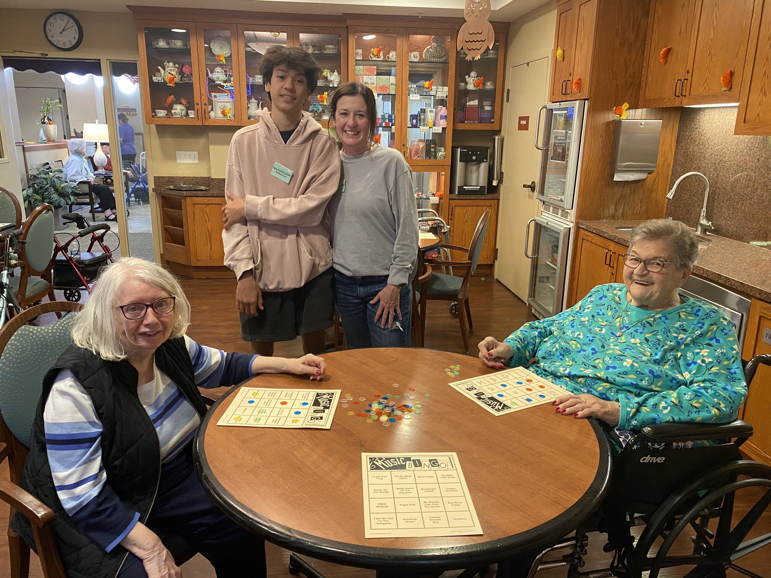 Four women, two seated at a table with bingo and tokens, and two standing behind them, in a cozy room with wooden cabinets and a kitchen area.