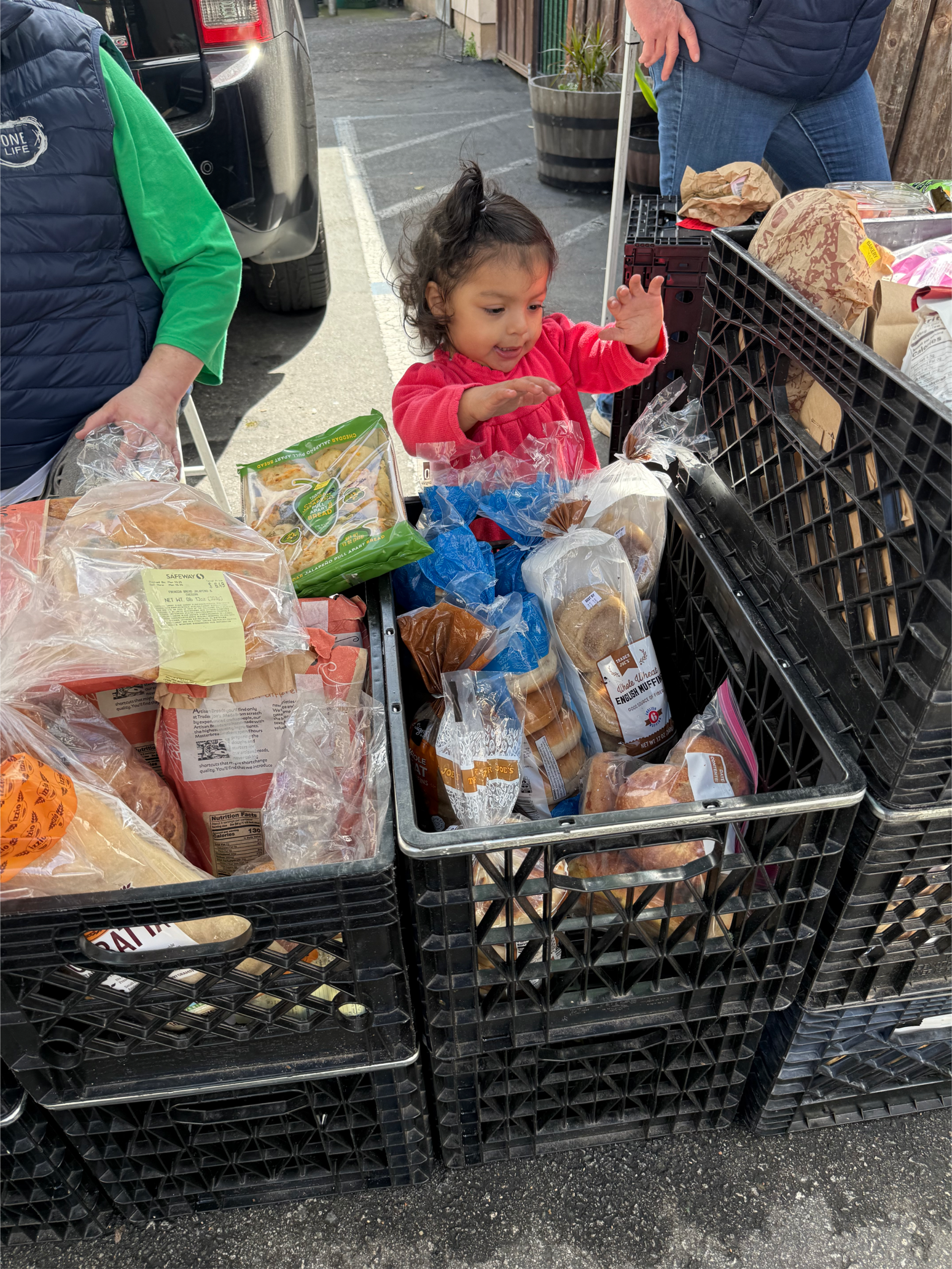 A young girl in a pink jacket shopping for bread and baked goods at an outdoor market, surrounded by plastic bags and crates filled with bread products, with adults nearby.