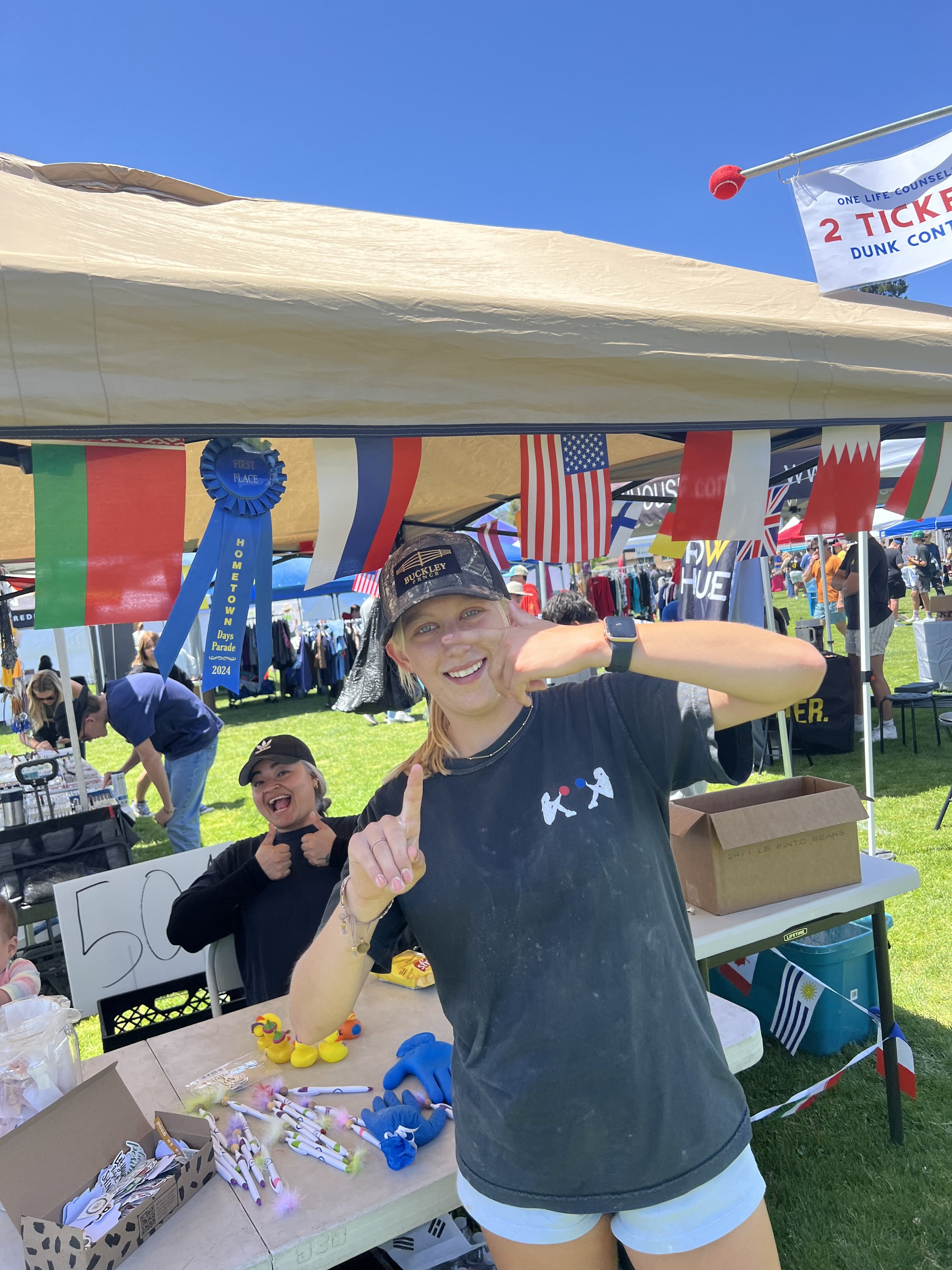 A young woman with blonde hair smiling and making a gesture with her hand at an outdoor fair, with a woman behind her smiling, under a tent decorated with international flags and a blue ribbon for first place.