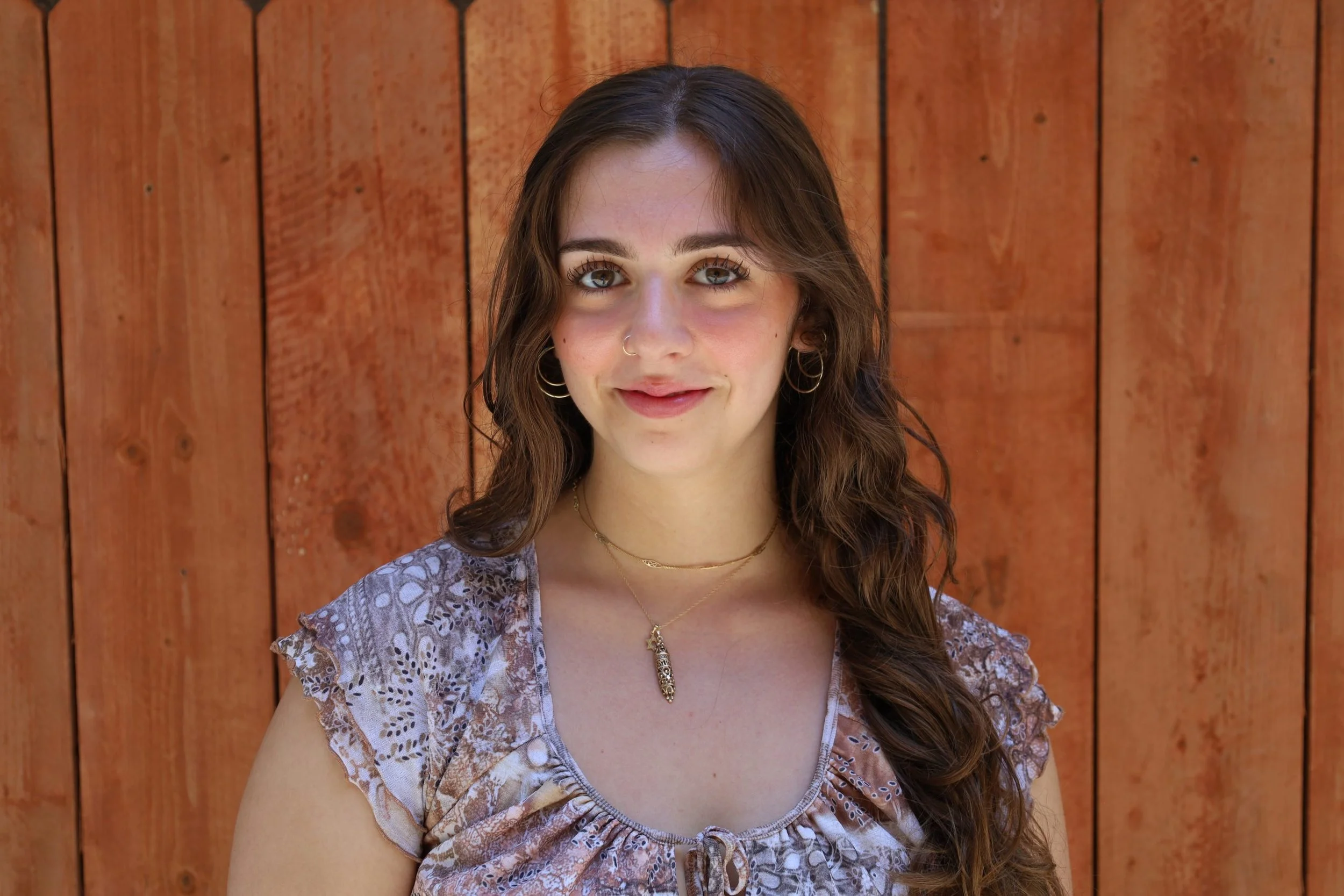 Close-up portrait of a young woman with long, wavy brown hair, wearing a patterned top and layered necklaces, standing in front of a wooden fence.
