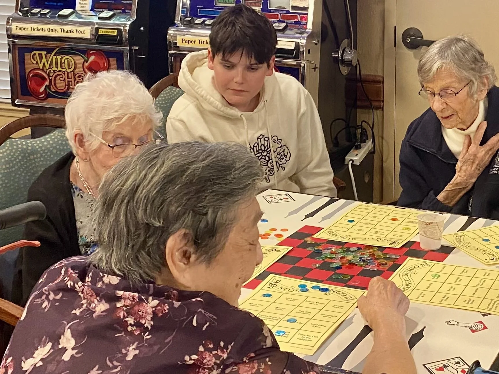 Group of elderly women and a young man playing bingo at a table, with bingo cards, colorful chips, and a small cup on the table in a gaming room with slot machines in the background.