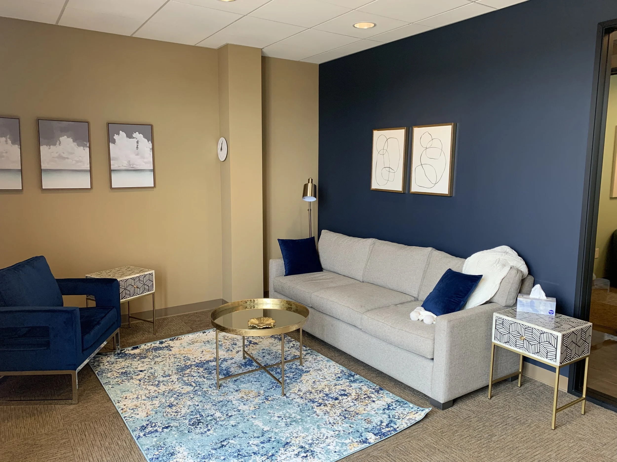 Living room with a beige wall and a navy accent wall, a light-colored sofa with blue pillows, a gold coffee table, a blue armchair, two patterned side tables, framed artwork, a wall clock, a floor lamp, a decorative rug, and a tissue box.