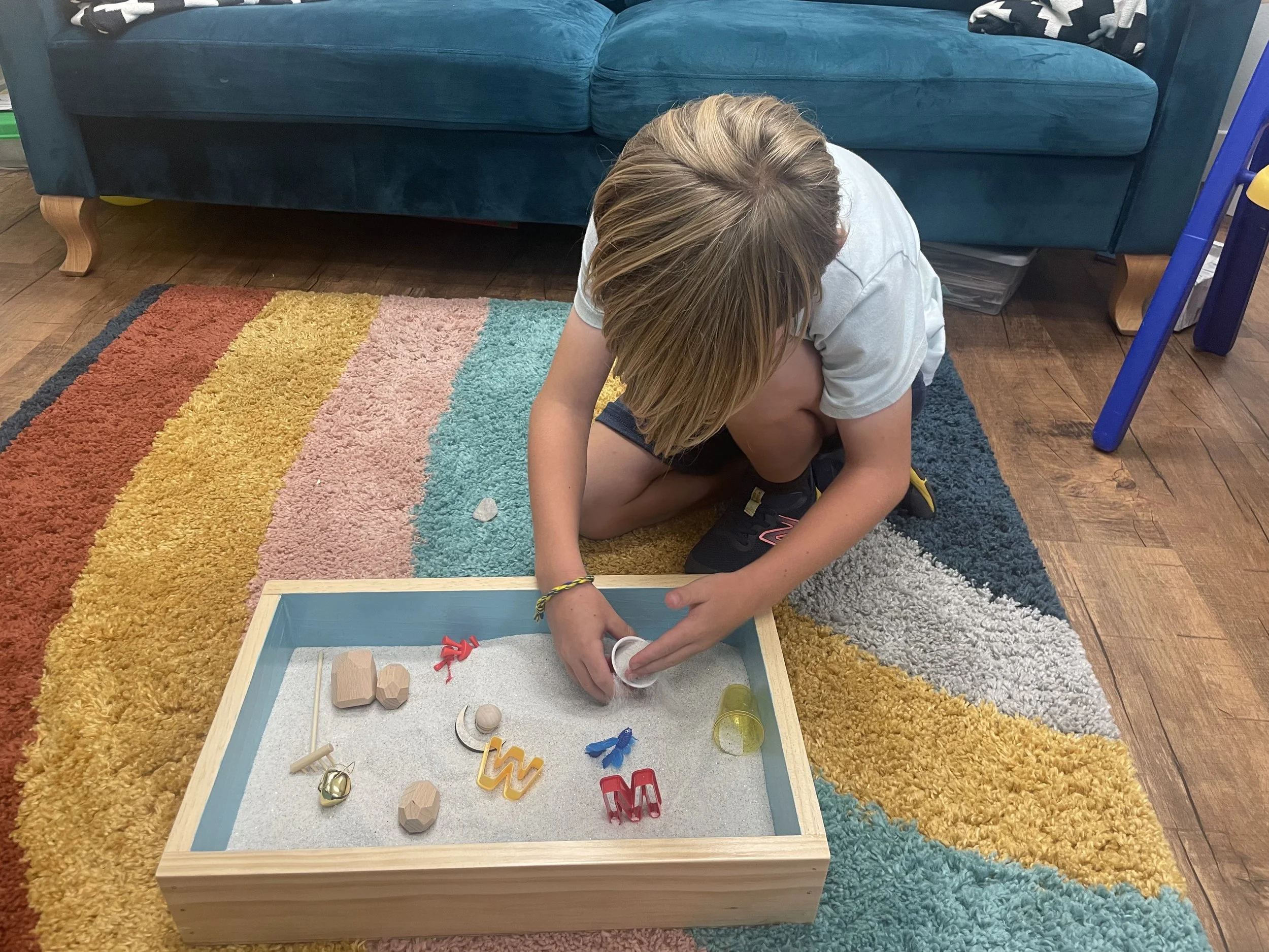 A child kneeling on a multicolored rug, playing with small wooden and plastic objects in a light wood box.
