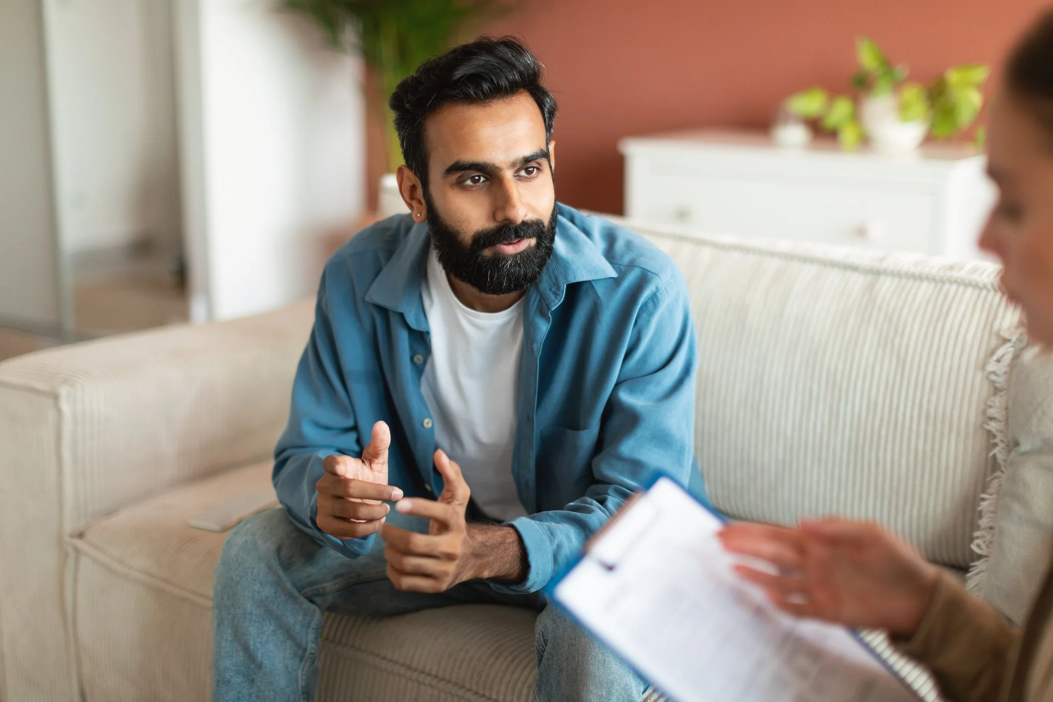 A man with dark hair, beard, wearing a blue shirt, sitting on a beige couch, talking to a woman with a clipboard, in a cozy room with potted plants and a white cabinet.