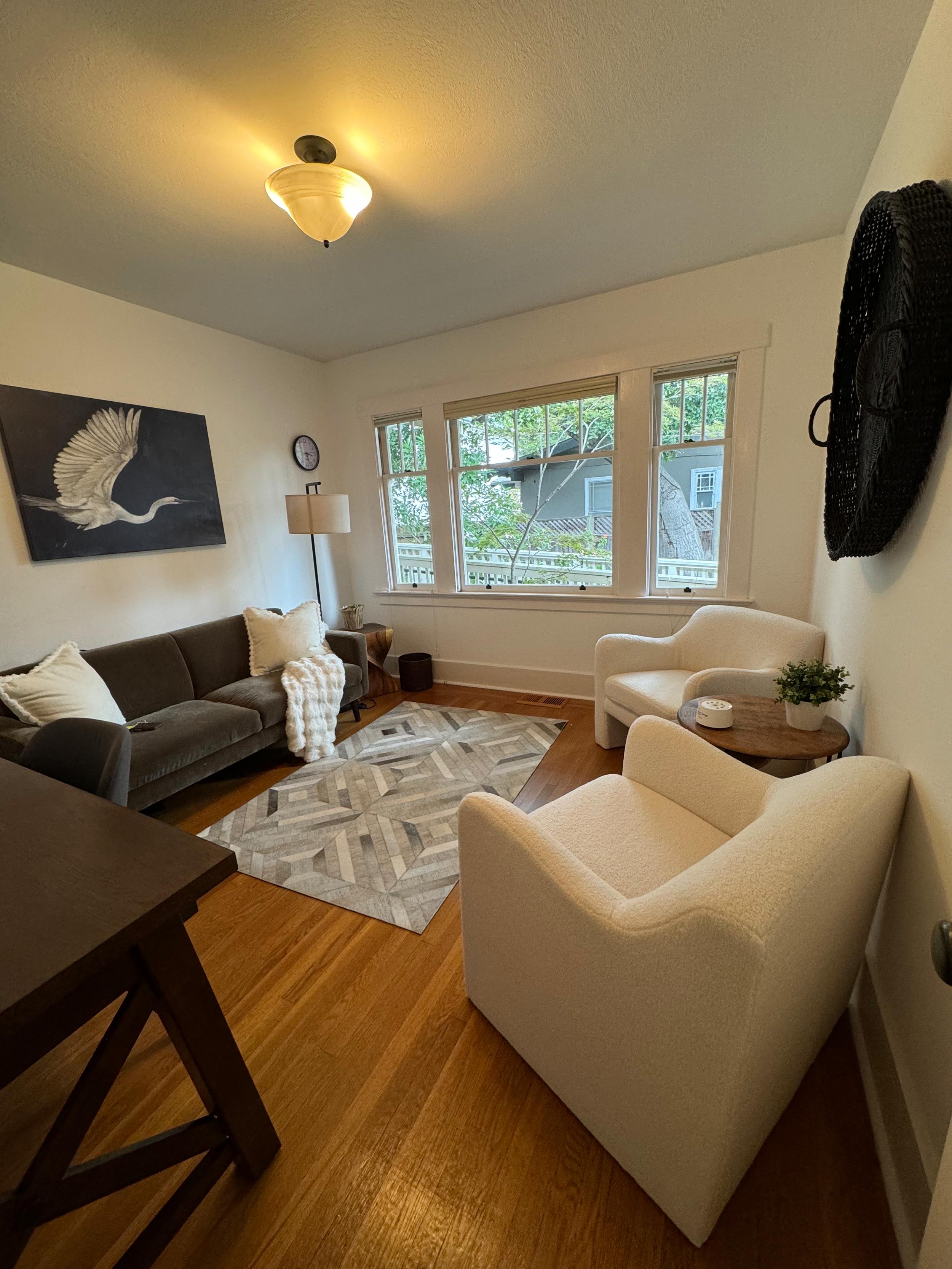 Living room with a gray sofa, white armchair, and a white chair, a patterned rug, a large window with greenery outside, and wall art of a flying bird.